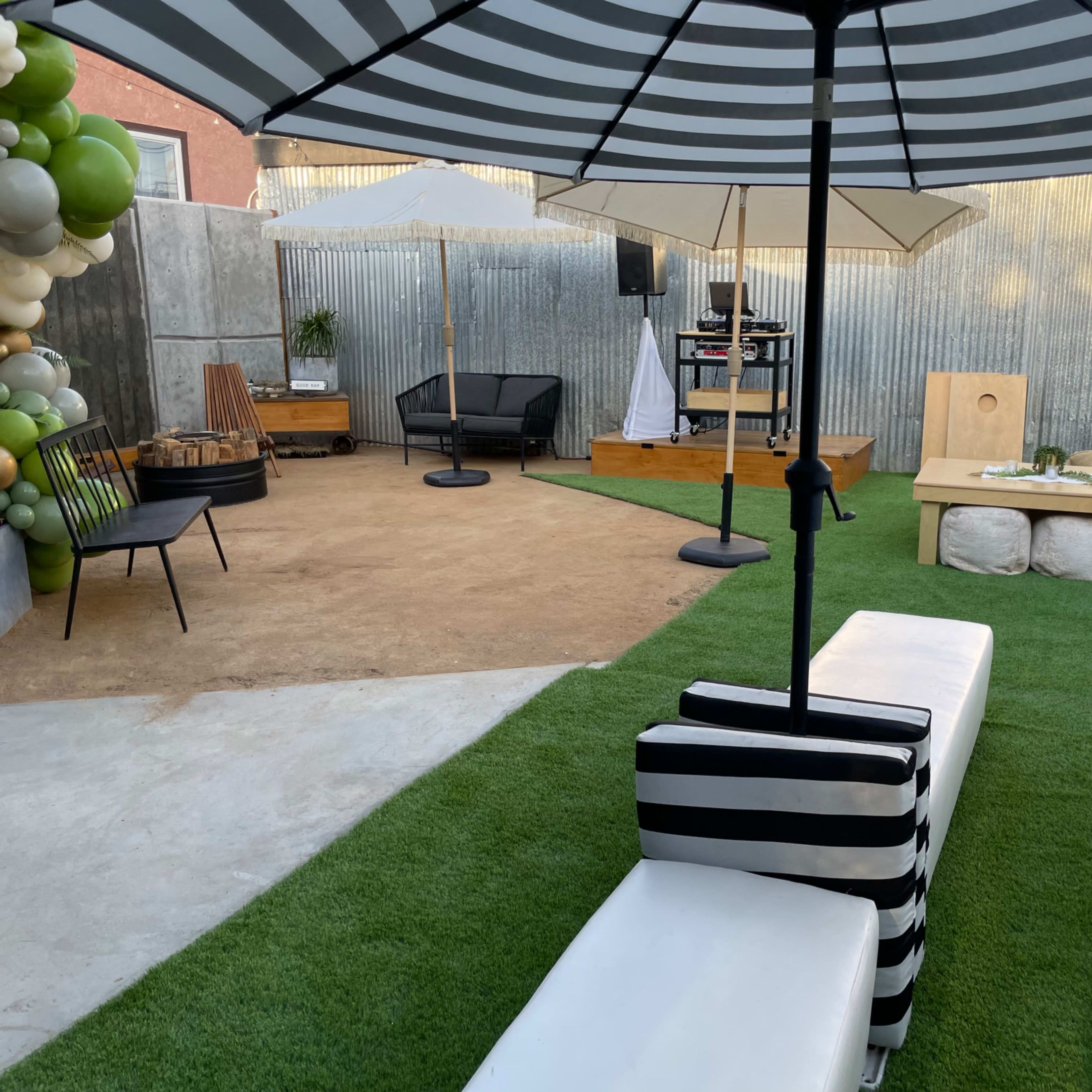 A landscaped outdoor area featuring seating, an umbrella, decorative elements, and a stage setup against a corrugated metal backdrop.