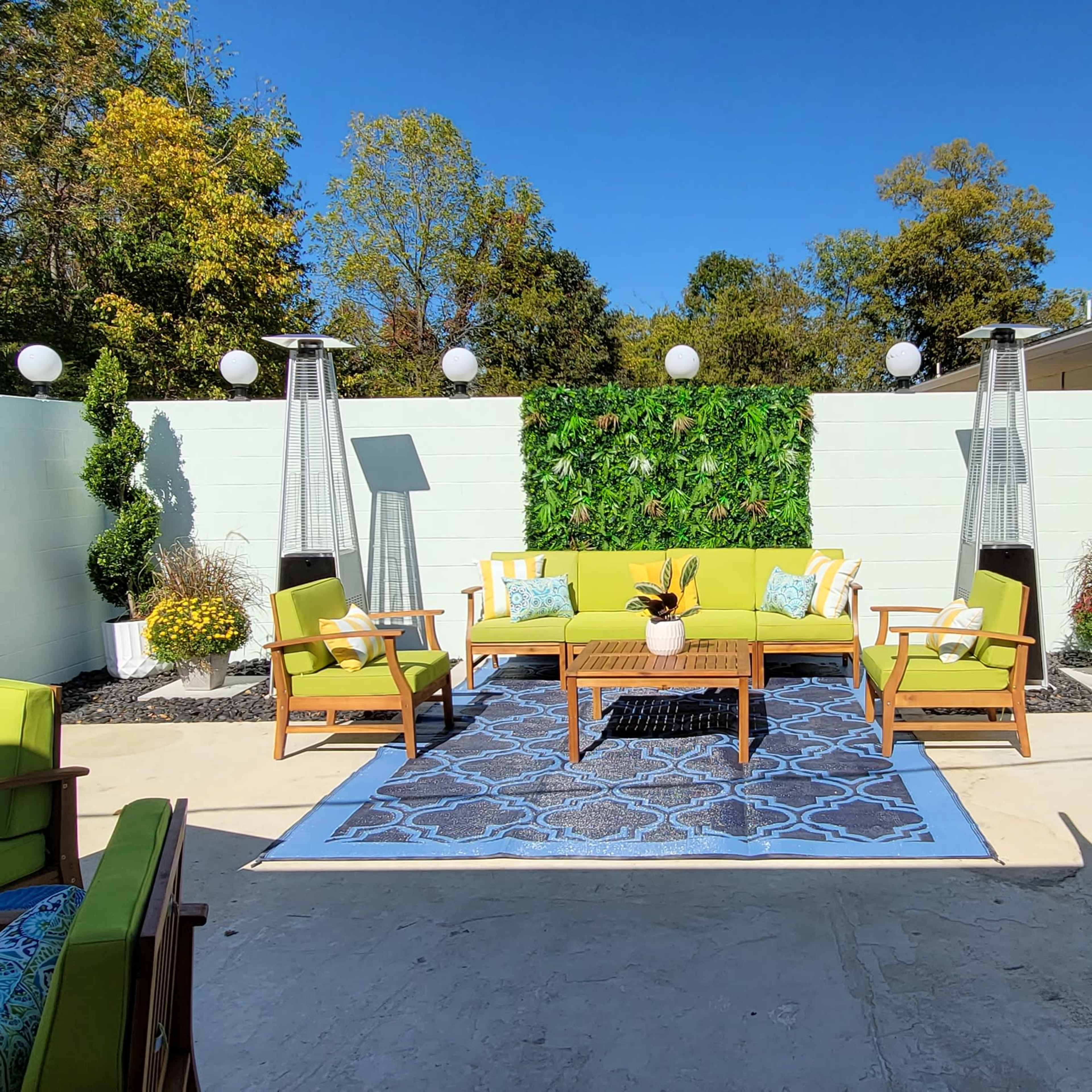 The outdoor space features a bright yellow sofa and chairs arranged around a patterned rug, flanked by decorative plants and lighting, with a patio dining set nearby.