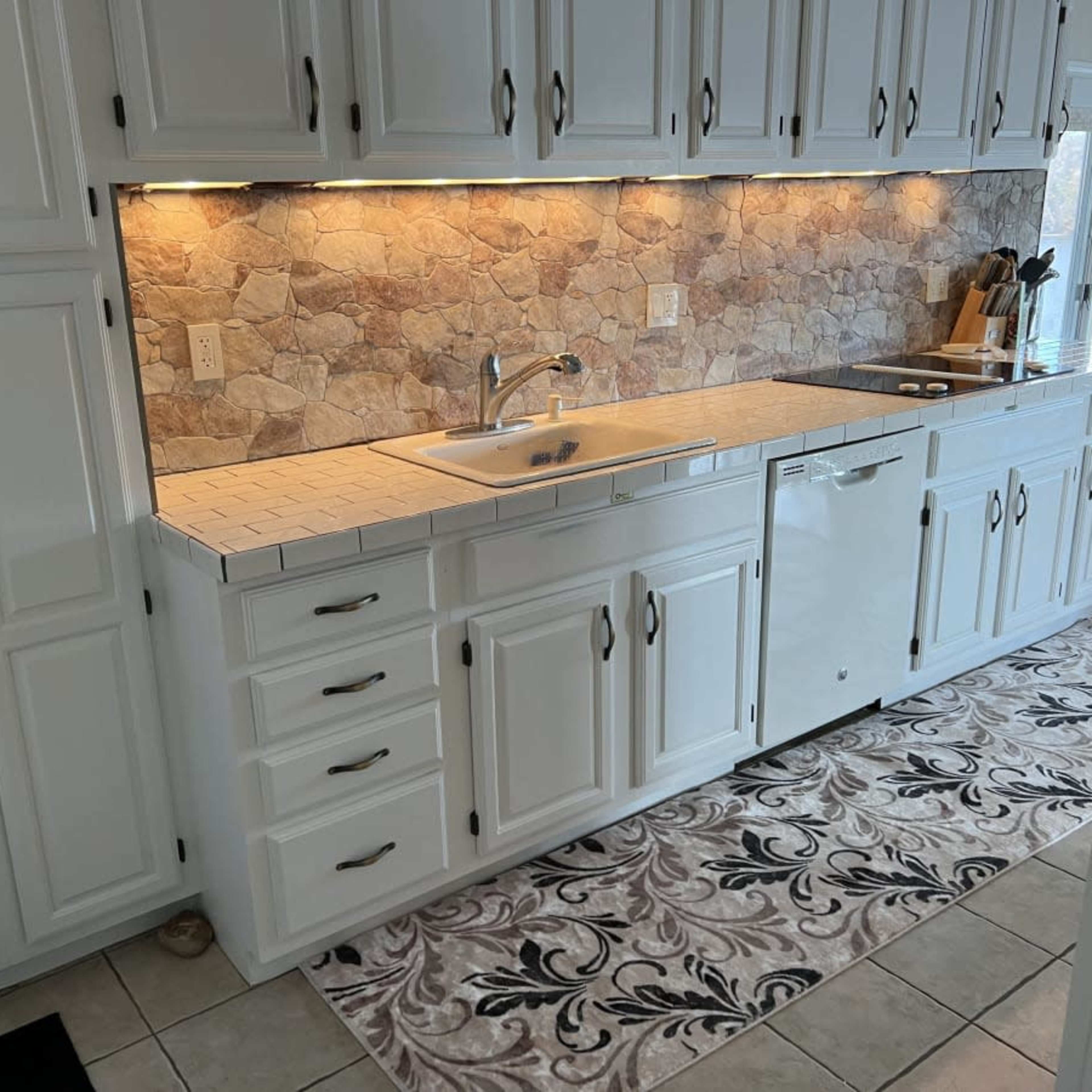 A kitchen with white cabinets, a stone backsplash, a sink, and a dishwashing machine, featuring a decorative rug on the floor.
