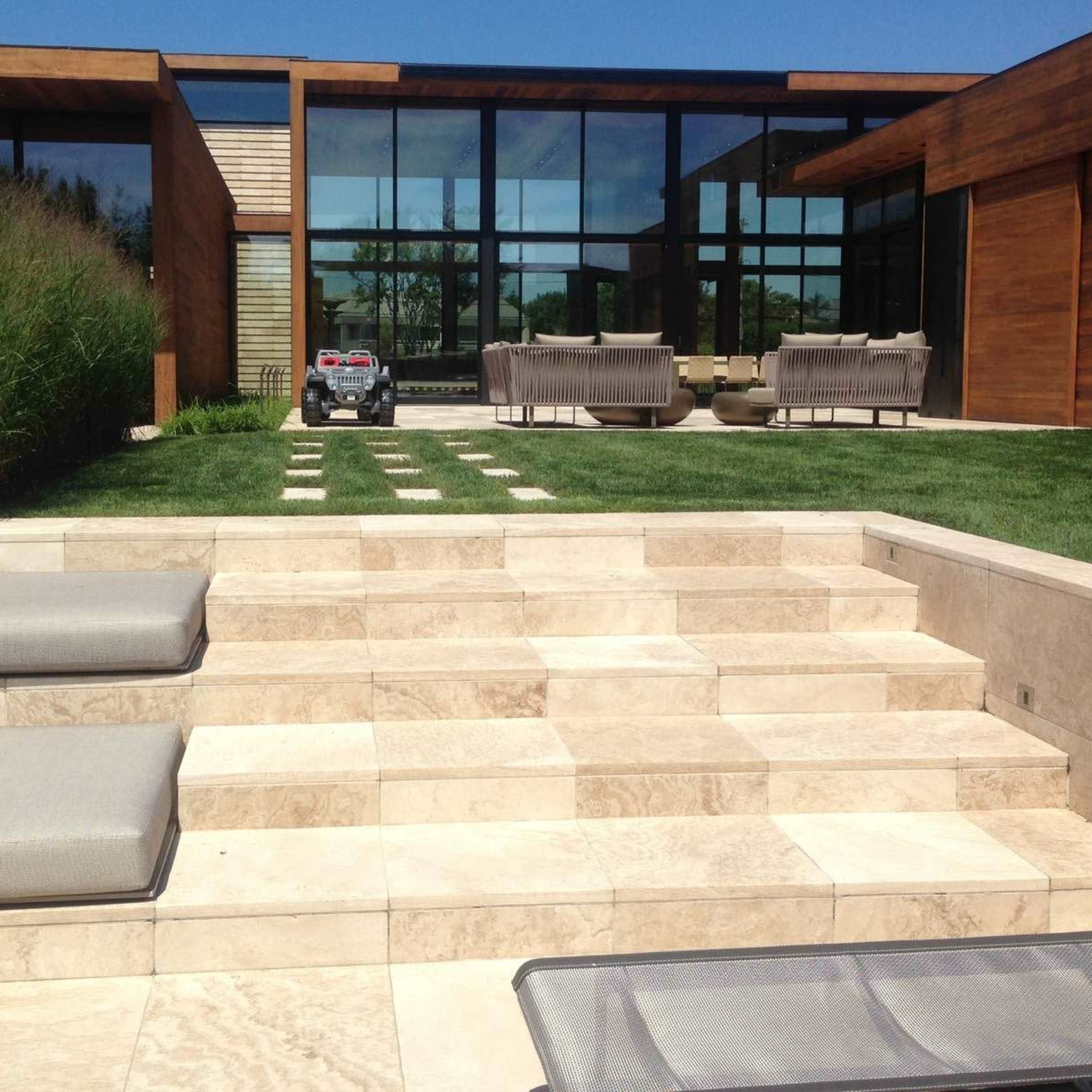 A modern backyard with stone steps leading up to a grassy area, surrounded by tall grasses and featuring a house with large glass windows.