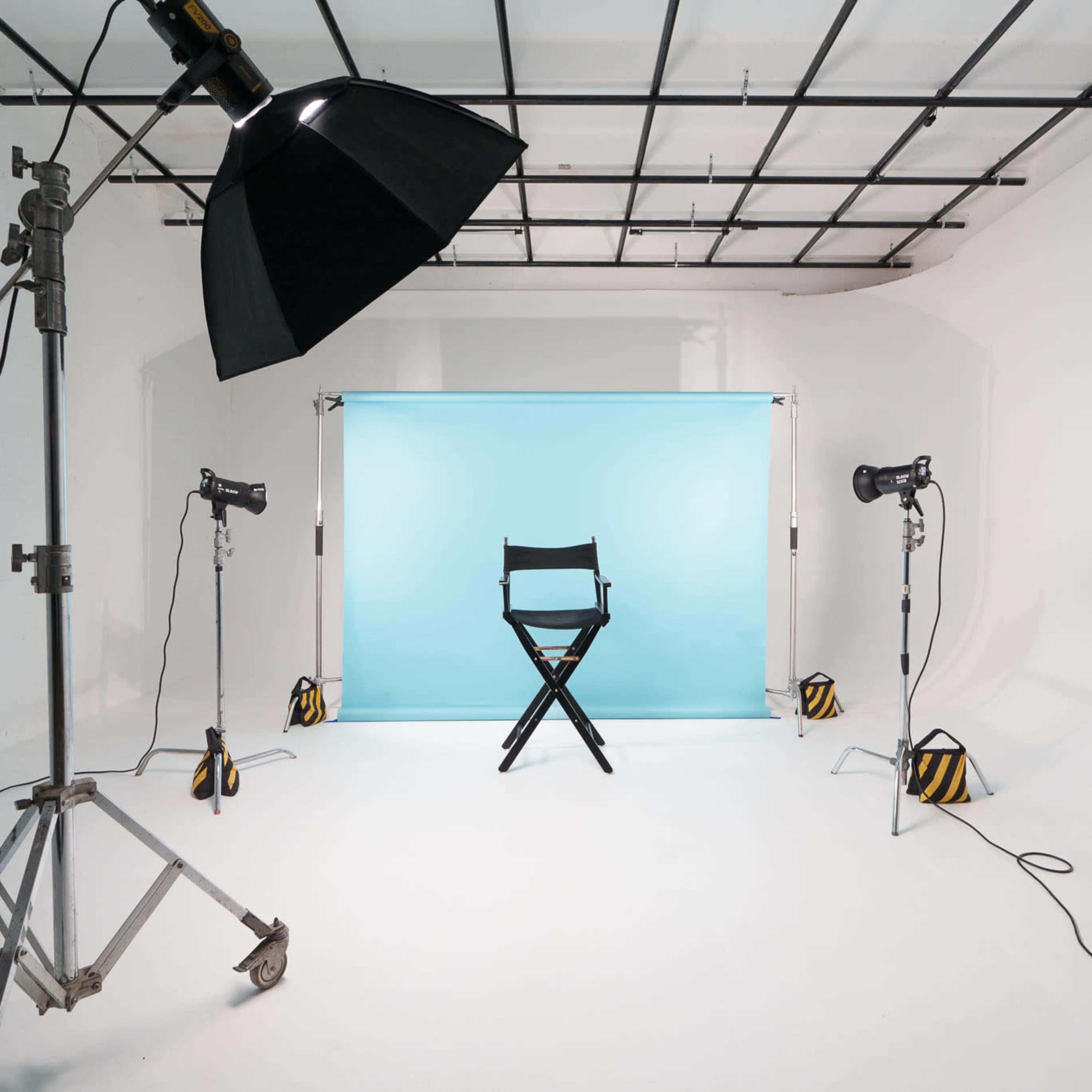 A photography studio features a black director's chair in front of a light blue backdrop, surrounded by studio lights and equipment.