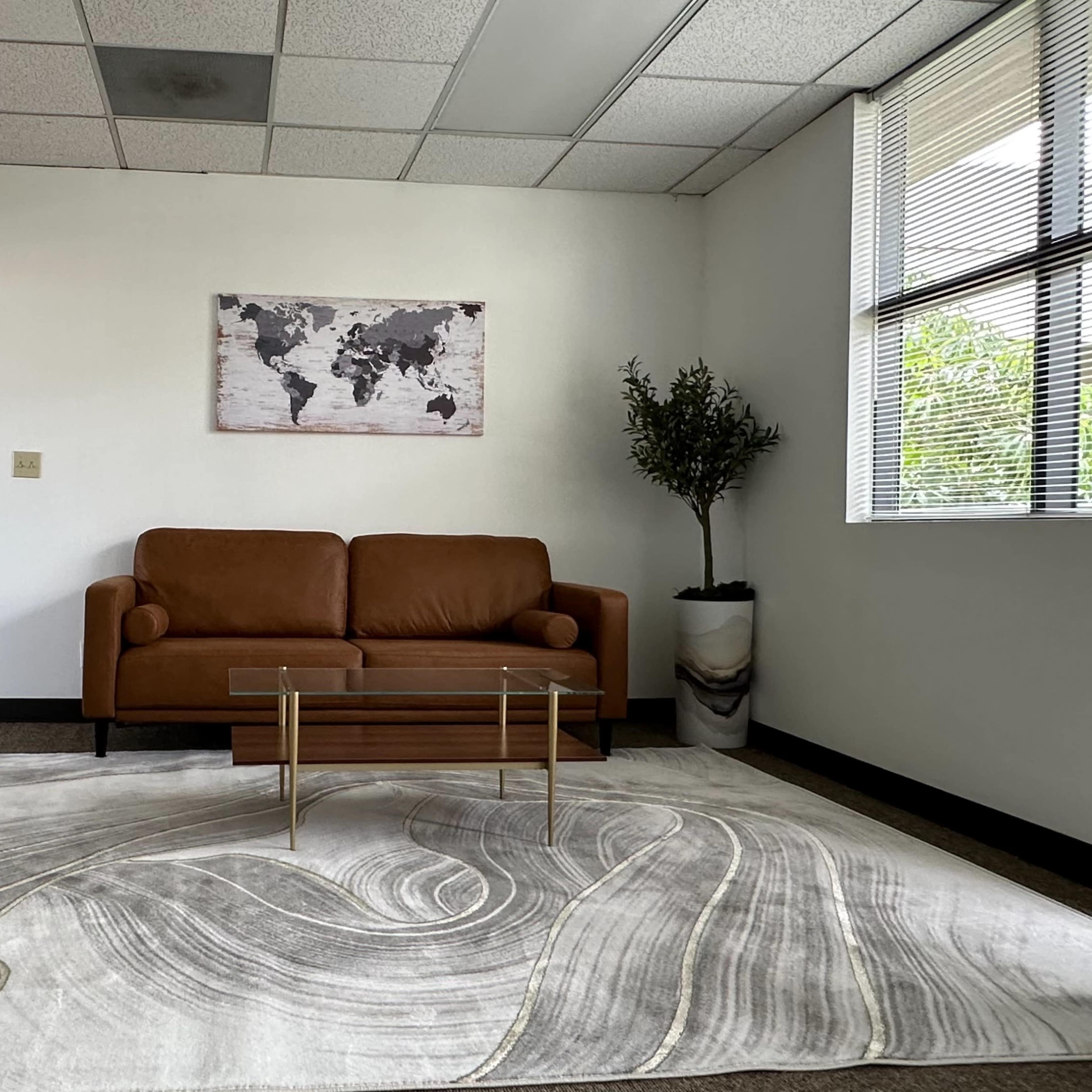 A modern office room features a brown sofa, a glass coffee table, and a world map on the wall, with a potted plant near a window.