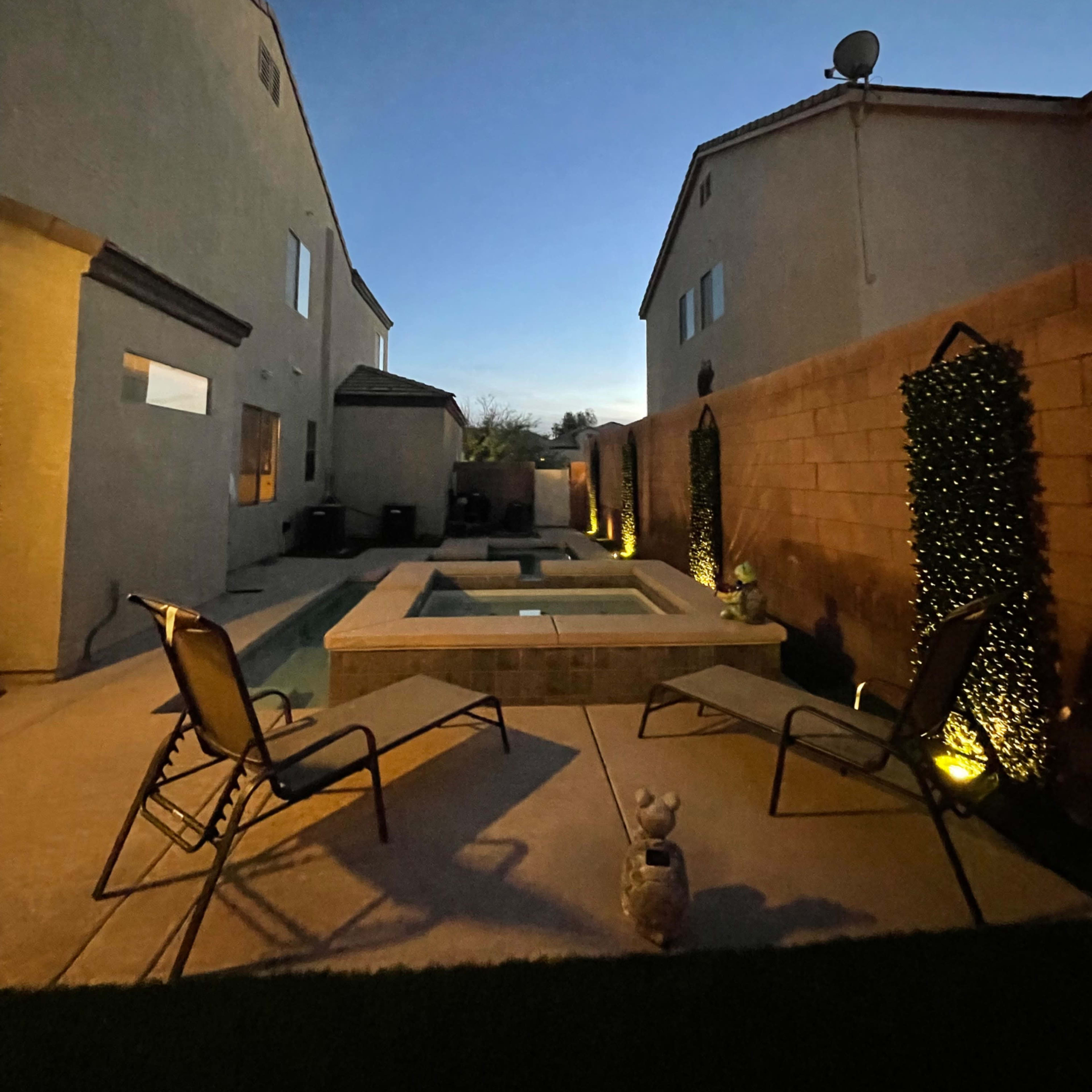 A backyard with a rectangular hot tub surrounded by two empty lounge chairs, flanked by low lighting and residential buildings under a twilight sky.