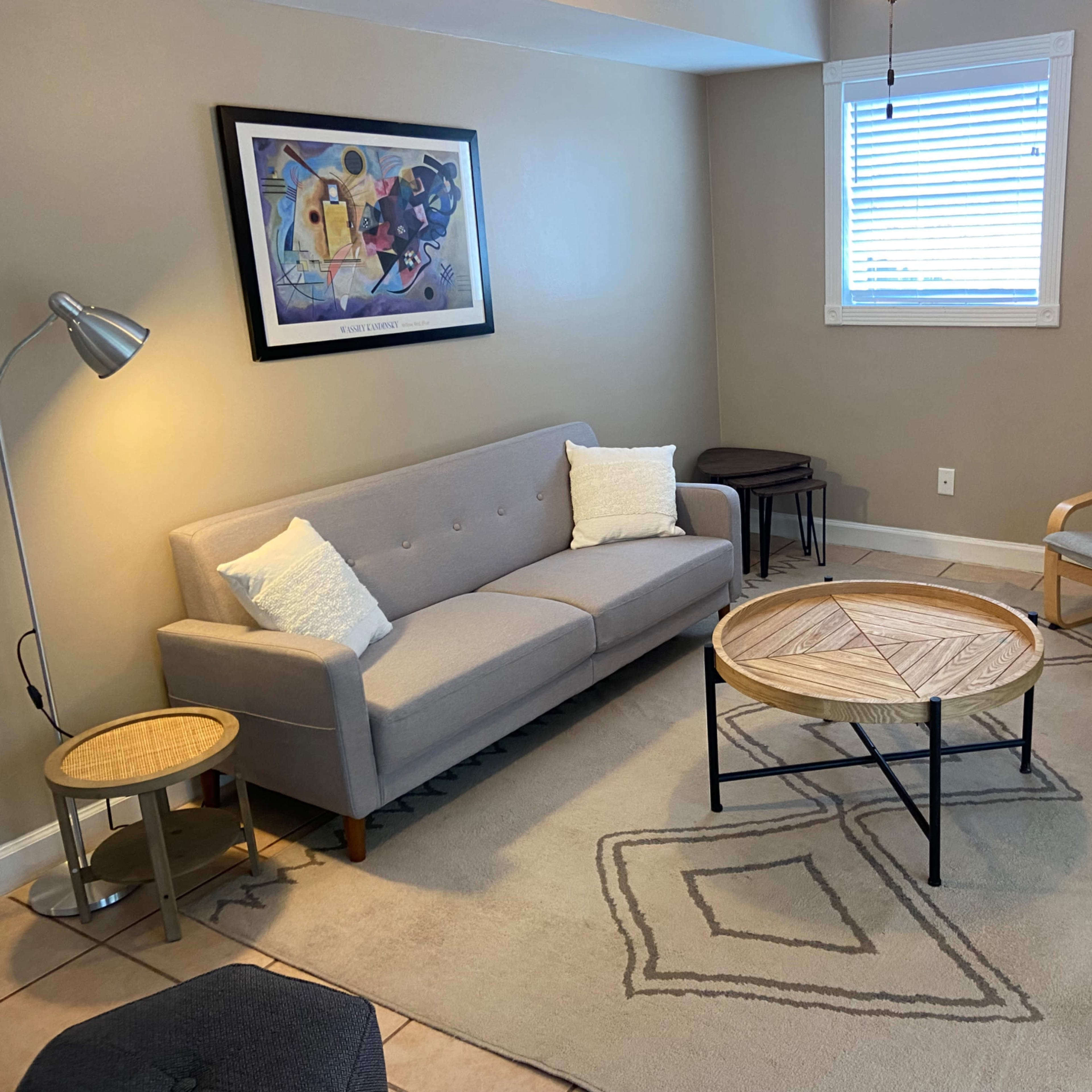 A cozy living room features a gray sofa, a wooden coffee table with a geometric design, a light-colored armchair, and a lamp, set against a neutral wall.
