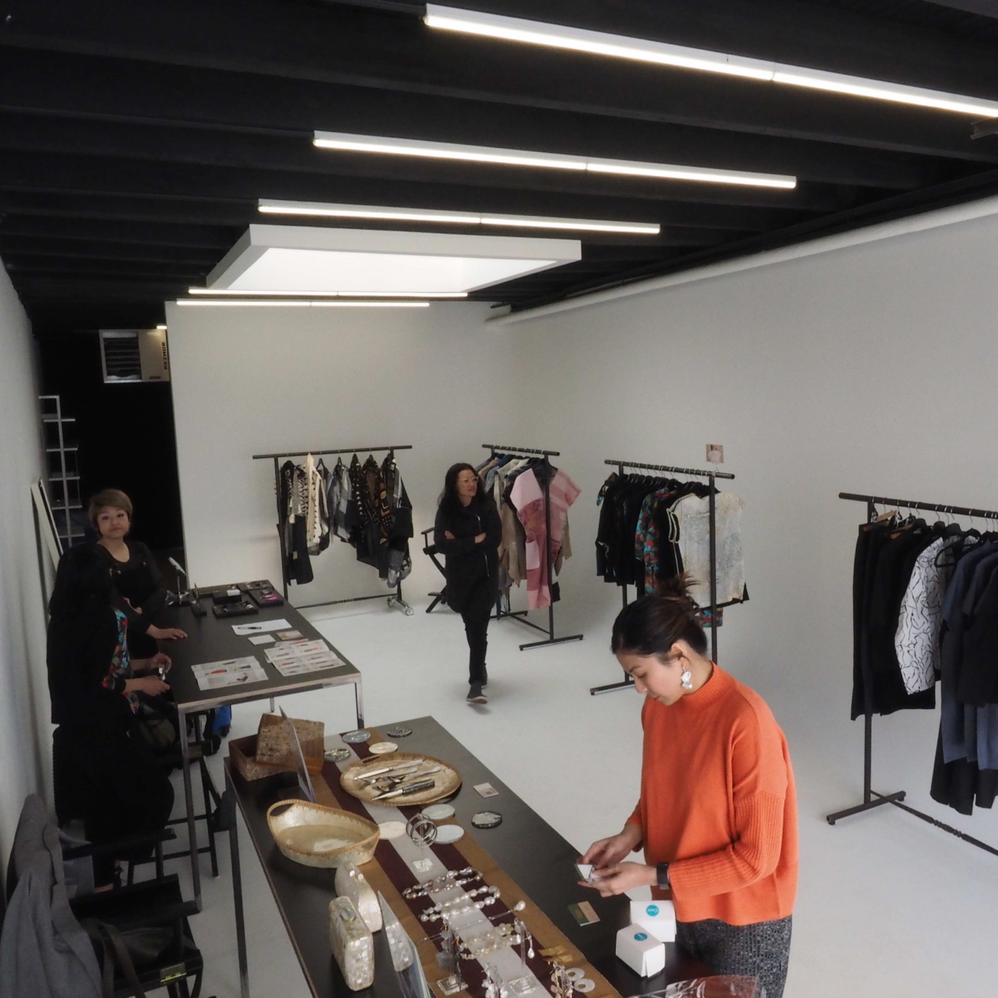 A jewelry and clothing boutique features a display table with accessories and multiple clothing racks against a minimalist white backdrop.