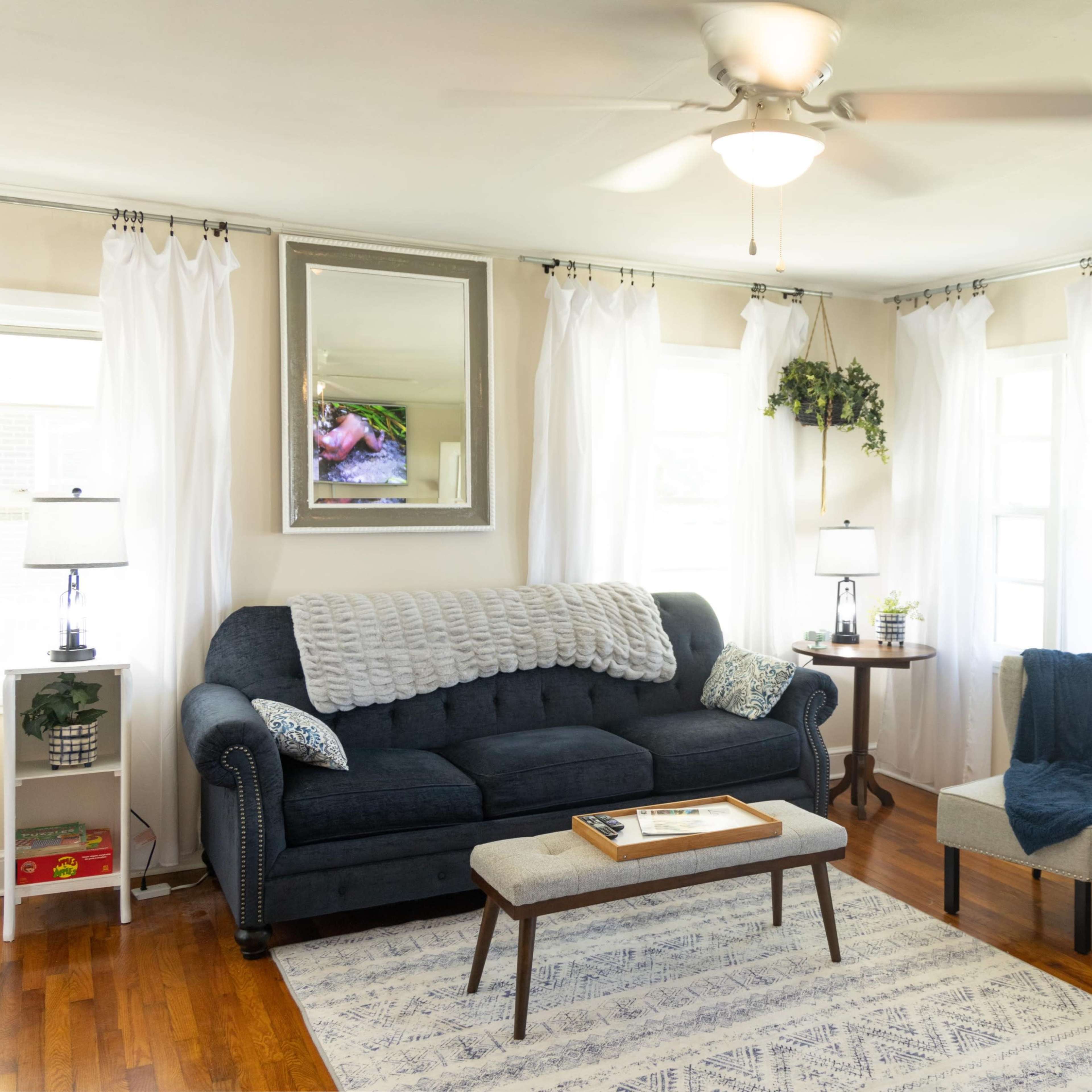 A bright living room features a blue sofa, two armchairs, a coffee table, and large windows adorned with white curtains.