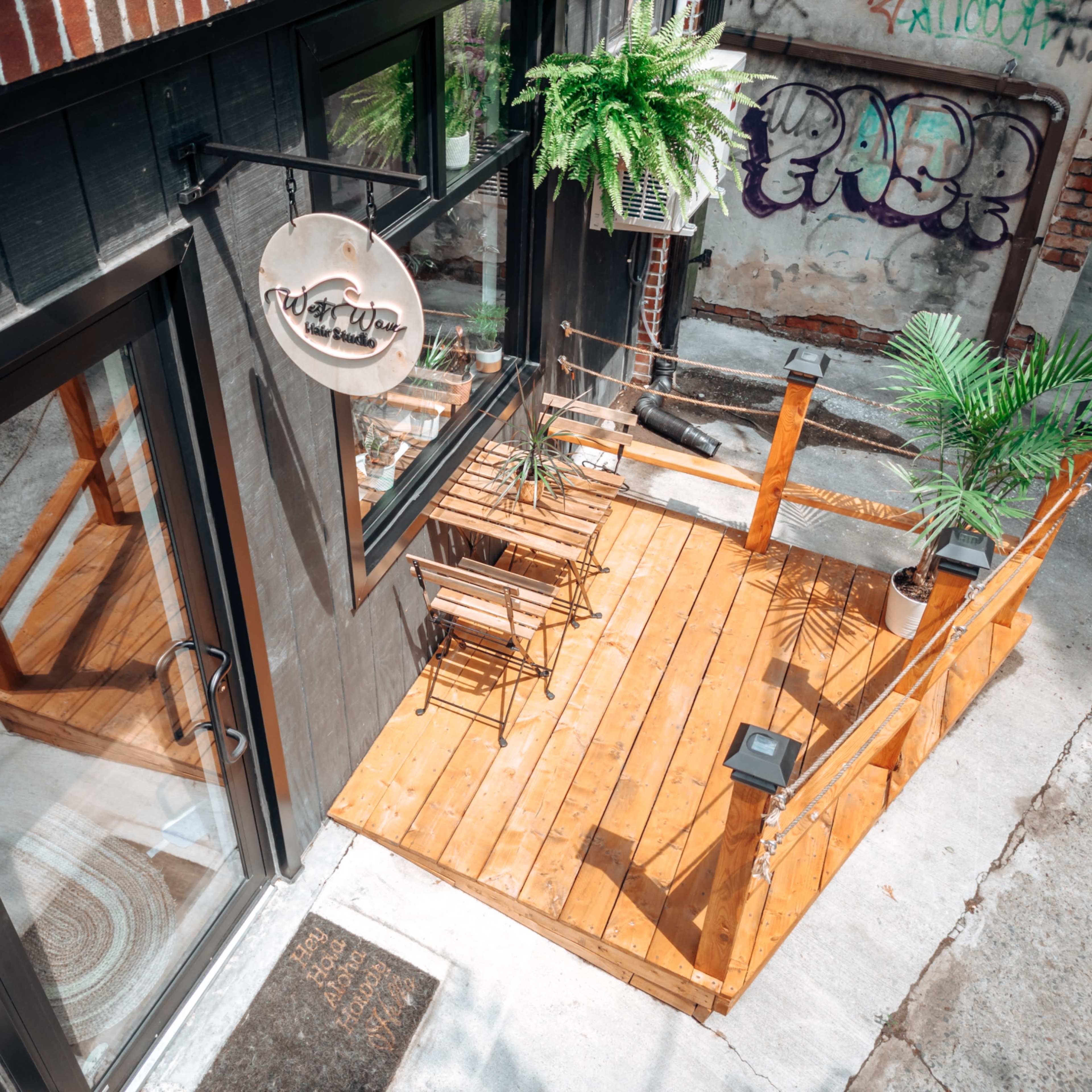 A small outdoor wooden deck with two chairs and a table, adjacent to a building with large windows and greenery, in an urban environment marked by graffiti on the walls.