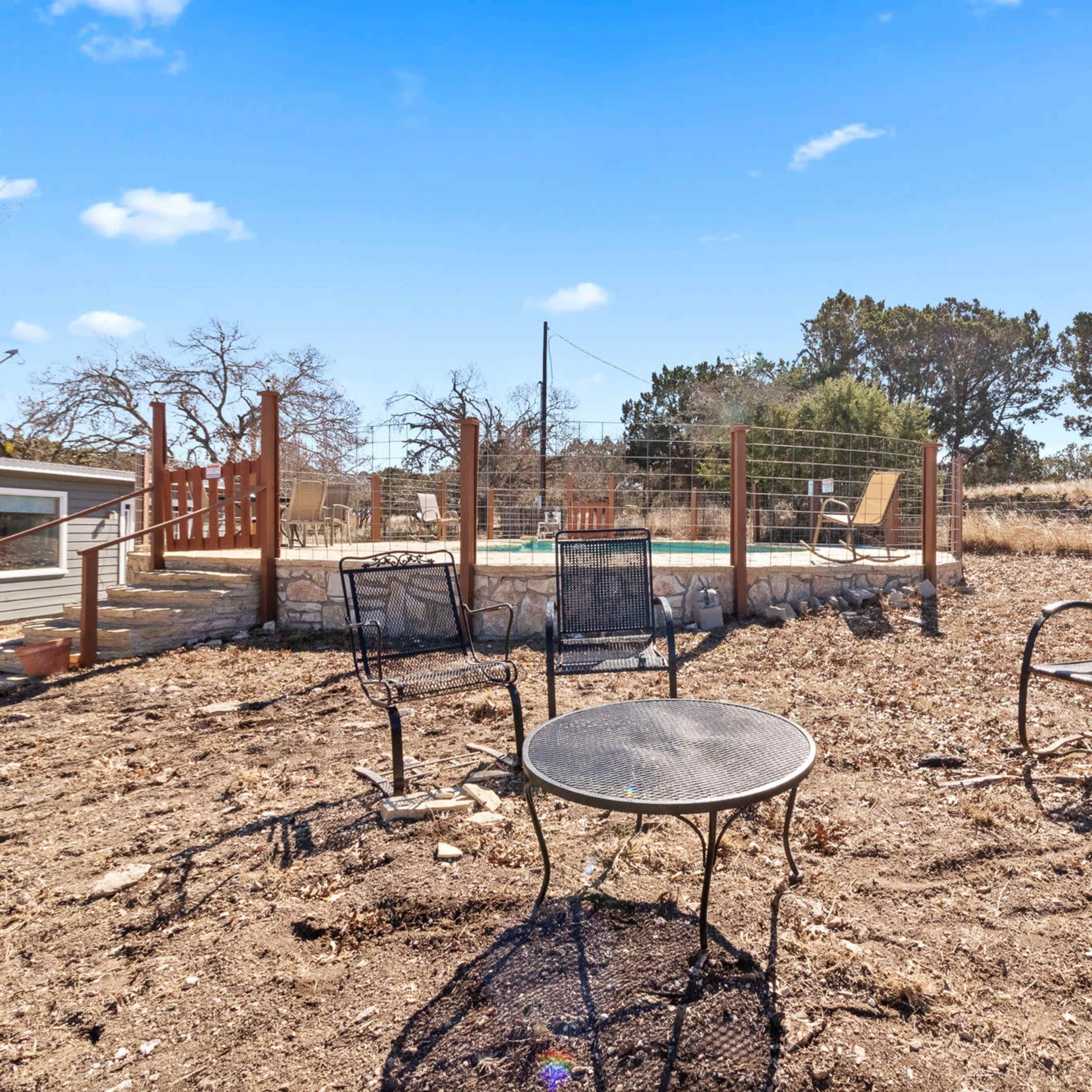 A barren outdoor area with several metal chairs and a table, overlooking a fenced pool surrounded by trees and a clear blue sky.