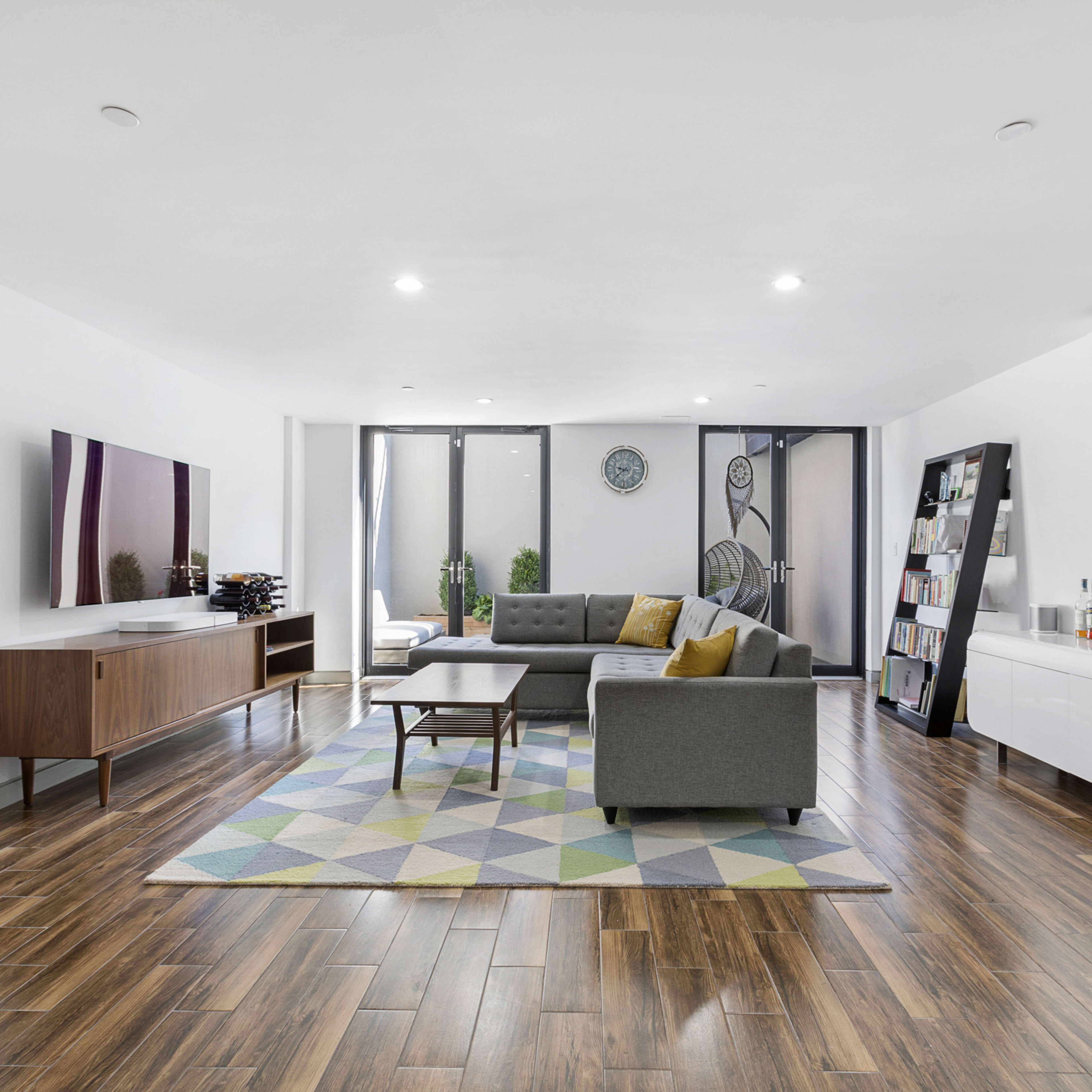 A modern living room with a sectional couch, coffee table, entertainment unit, and a bar cart, set against a backdrop of large windows and a geometric patterned rug.