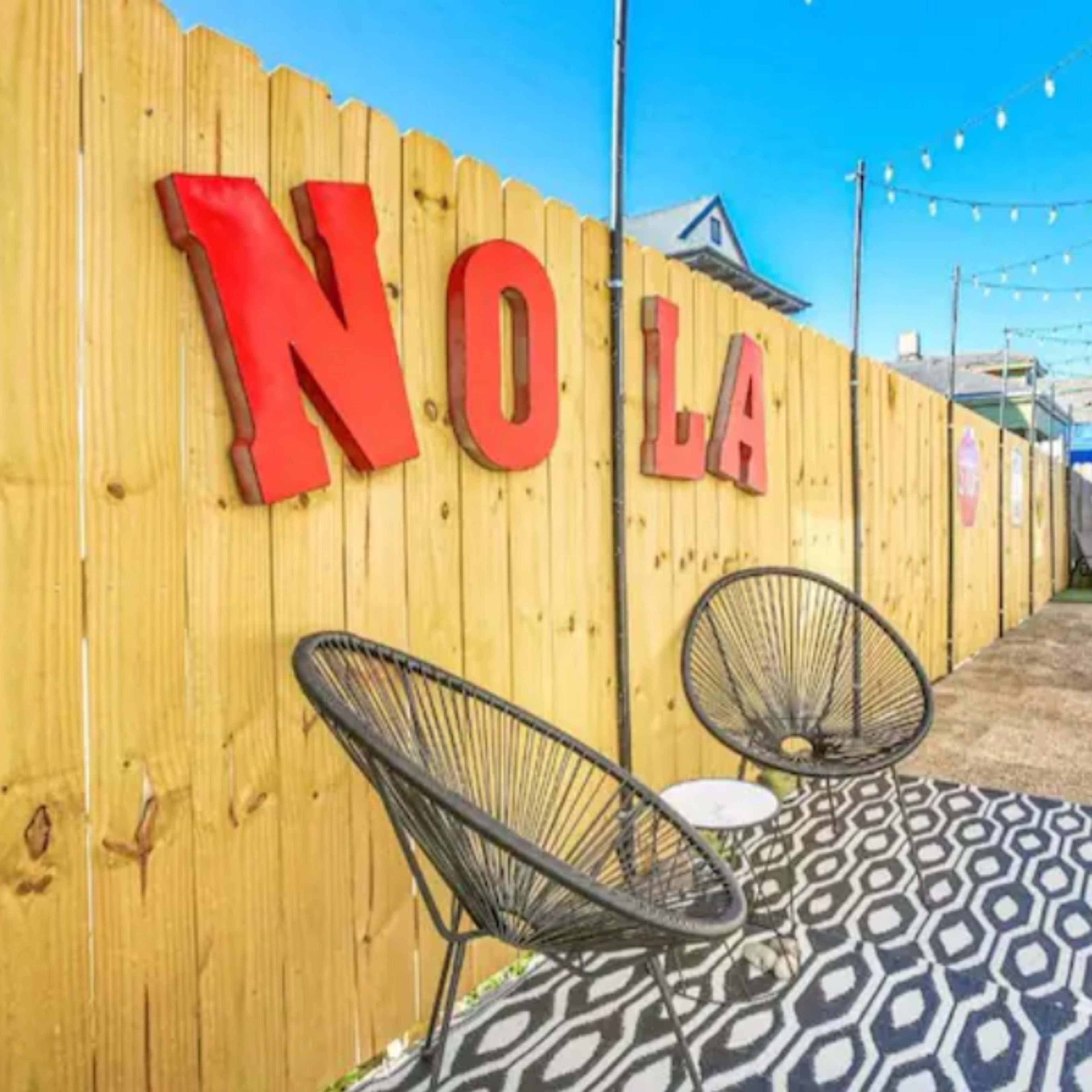 A narrow outdoor space features a wooden fence with the letters "NO LA," string lights above, and two black chairs on a patterned floor.