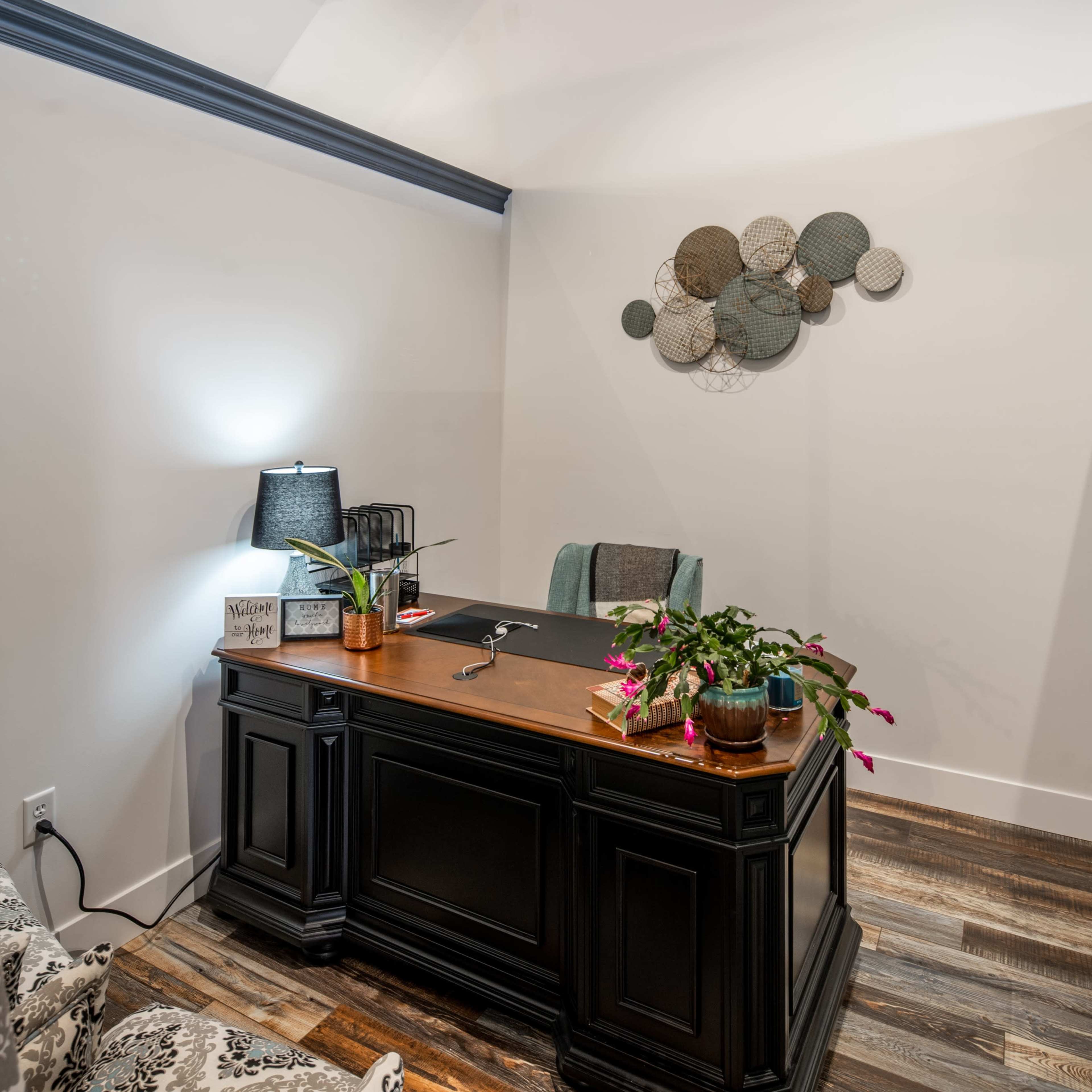 A modern office space features a dark desk, a lamp, and decorative wall art, with a plant on the desk and a cozy seating area nearby.