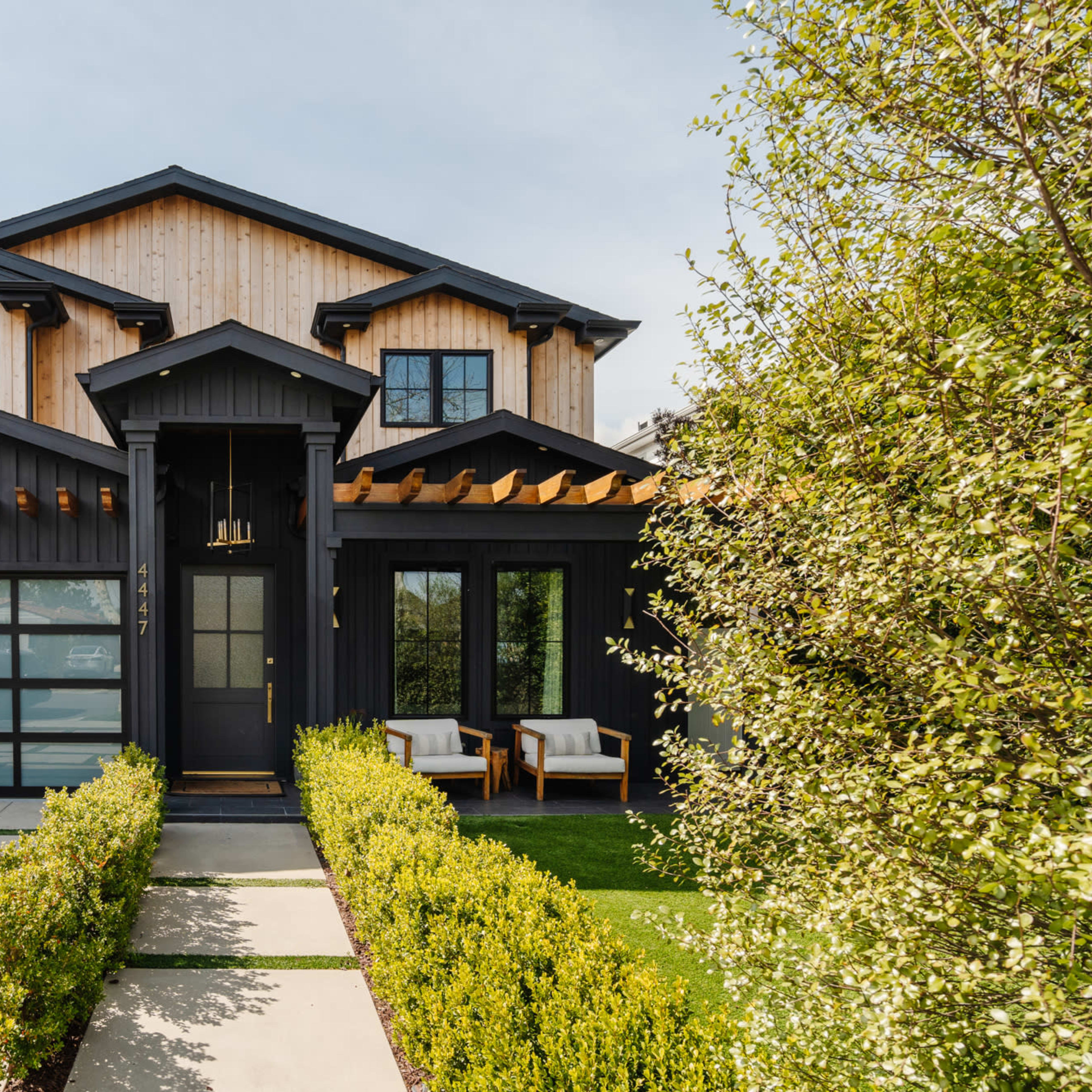 A modern black and wood house with large windows, a front porch, and a neatly landscaped pathway lined with green hedges.