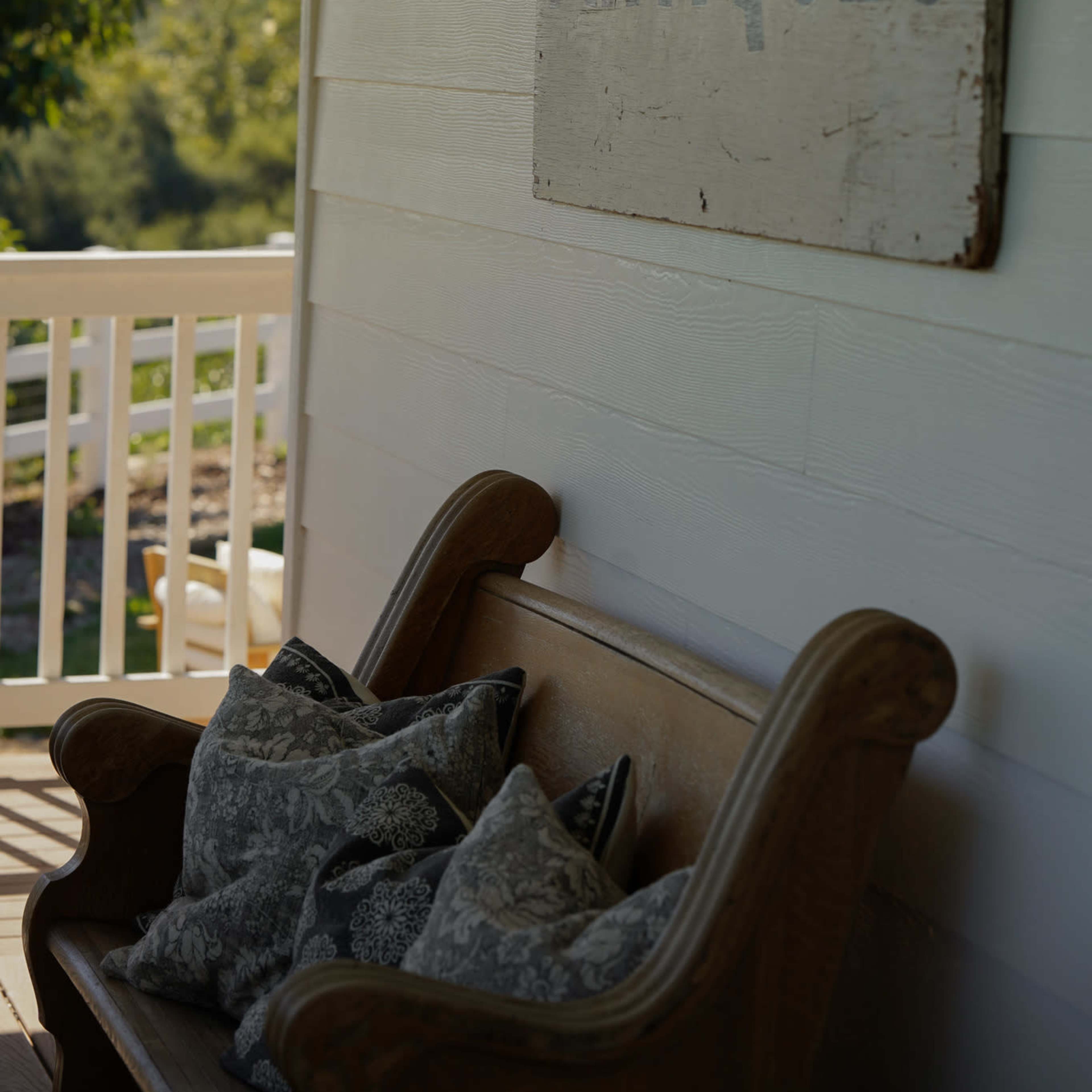 A wooden bench with decorative pillows beneath an "ANTIQUE" sign on a white wall, with greenery visible in the background.