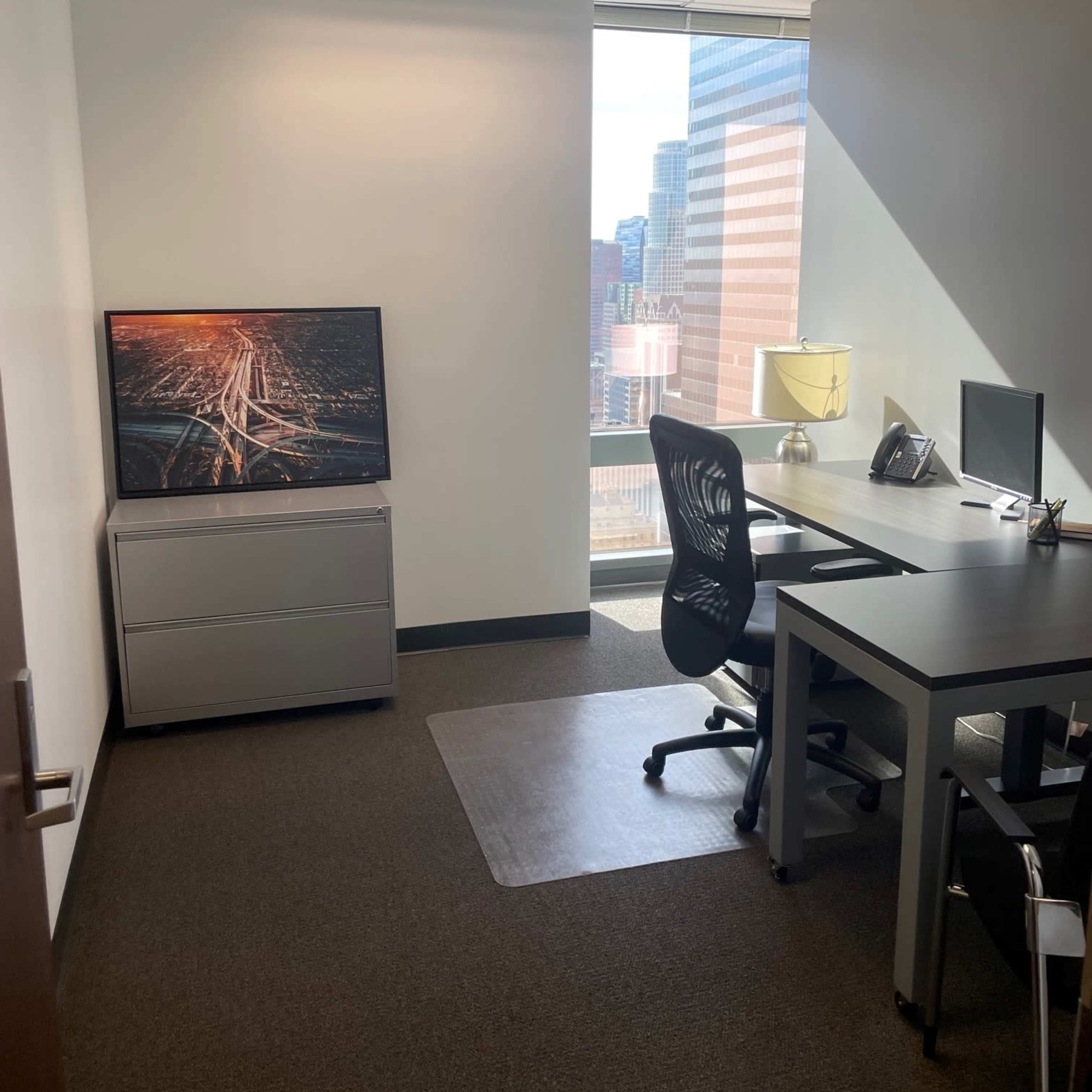 A modern office space featuring a desk with a computer, a chair, a television on a stand, and large windows overlooking a city skyline.