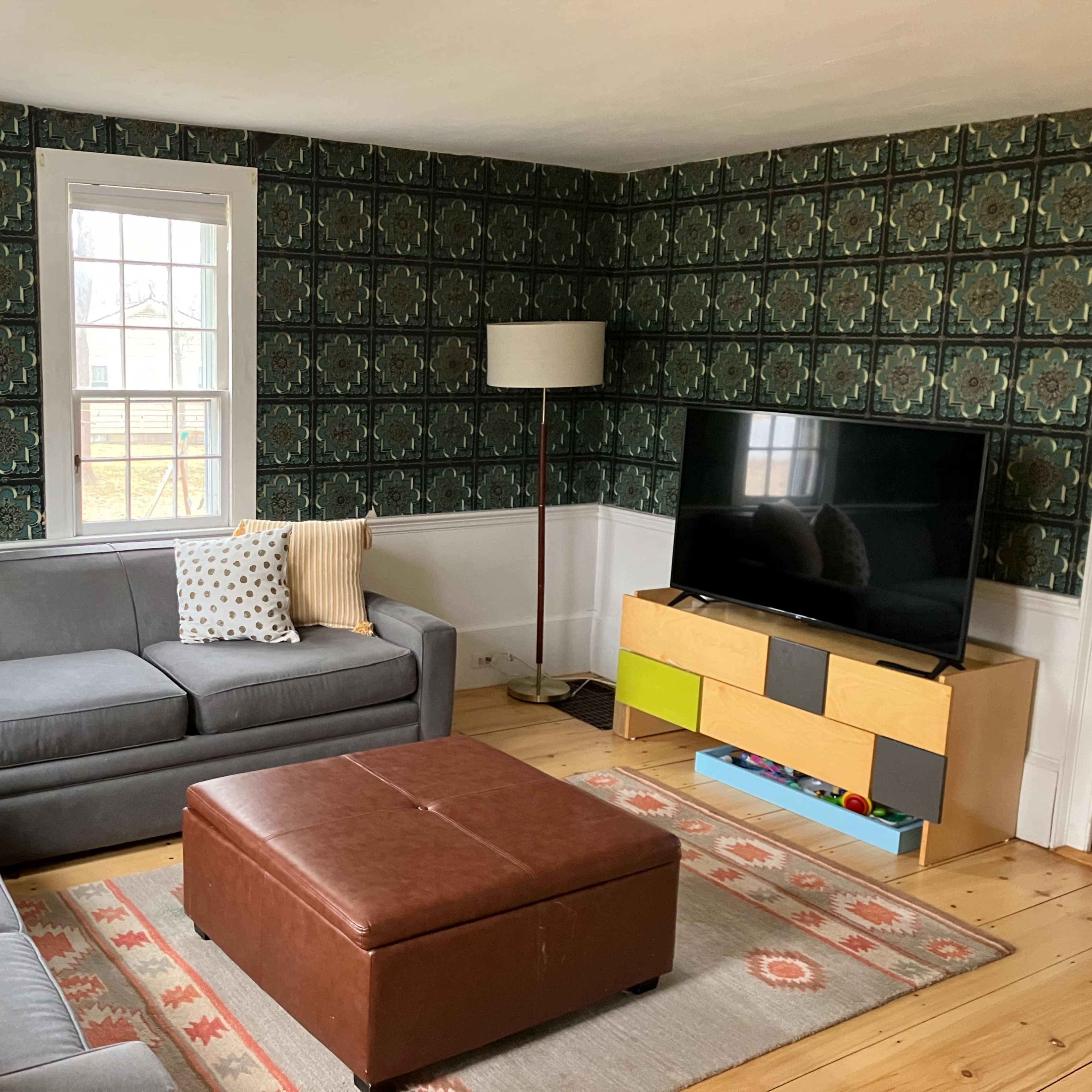 A living room features a gray sectional sofa, a brown ottoman, and a television on a low cabinet against a patterned wallpapered wall.