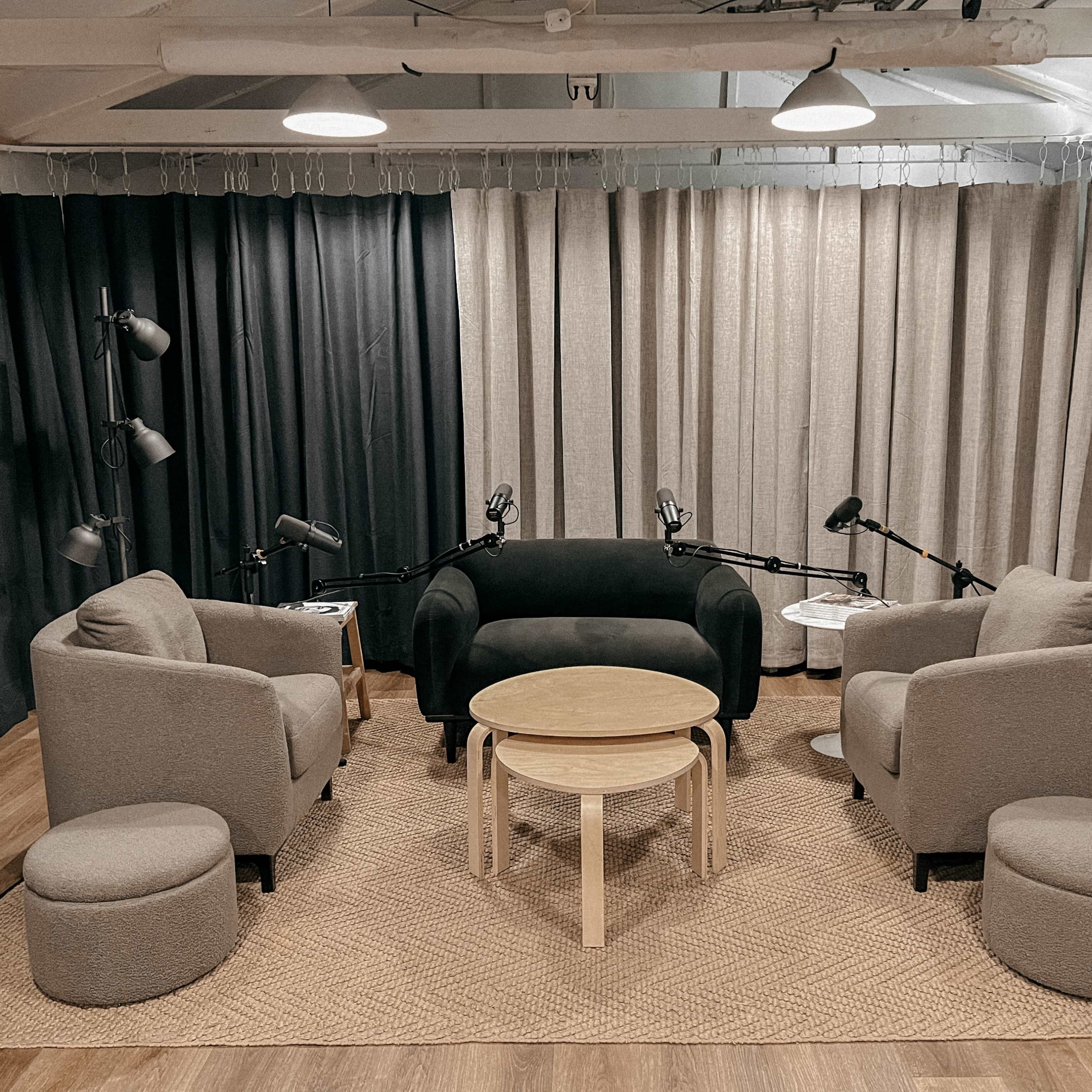 A cozy podcast studio features a central table surrounded by four armchairs and soft seating, with lamps and microphones positioned nearby.