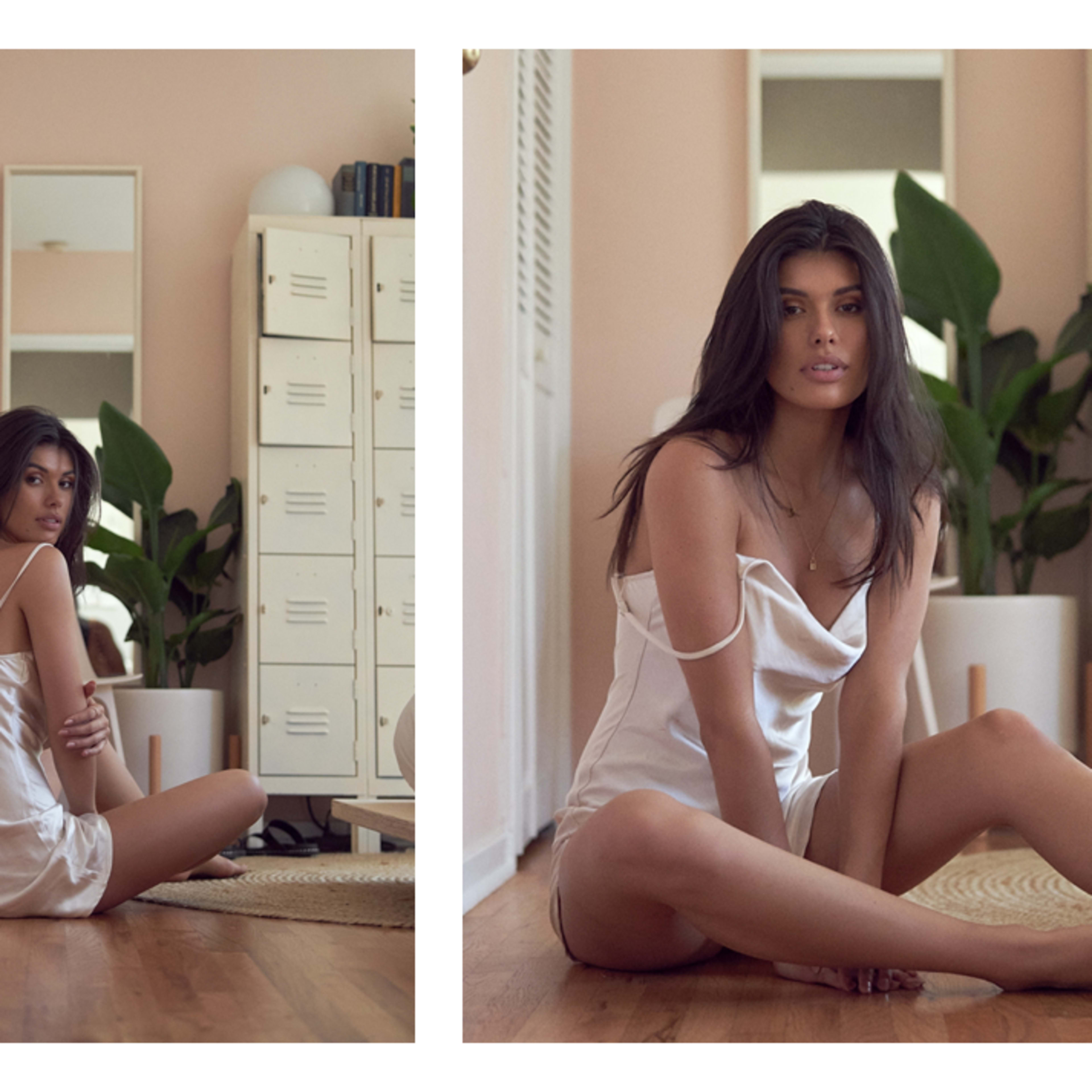 A woman sits on a wooden floor in a well-lit room, surrounded by plants and mirrors, wearing a light-colored outfit.