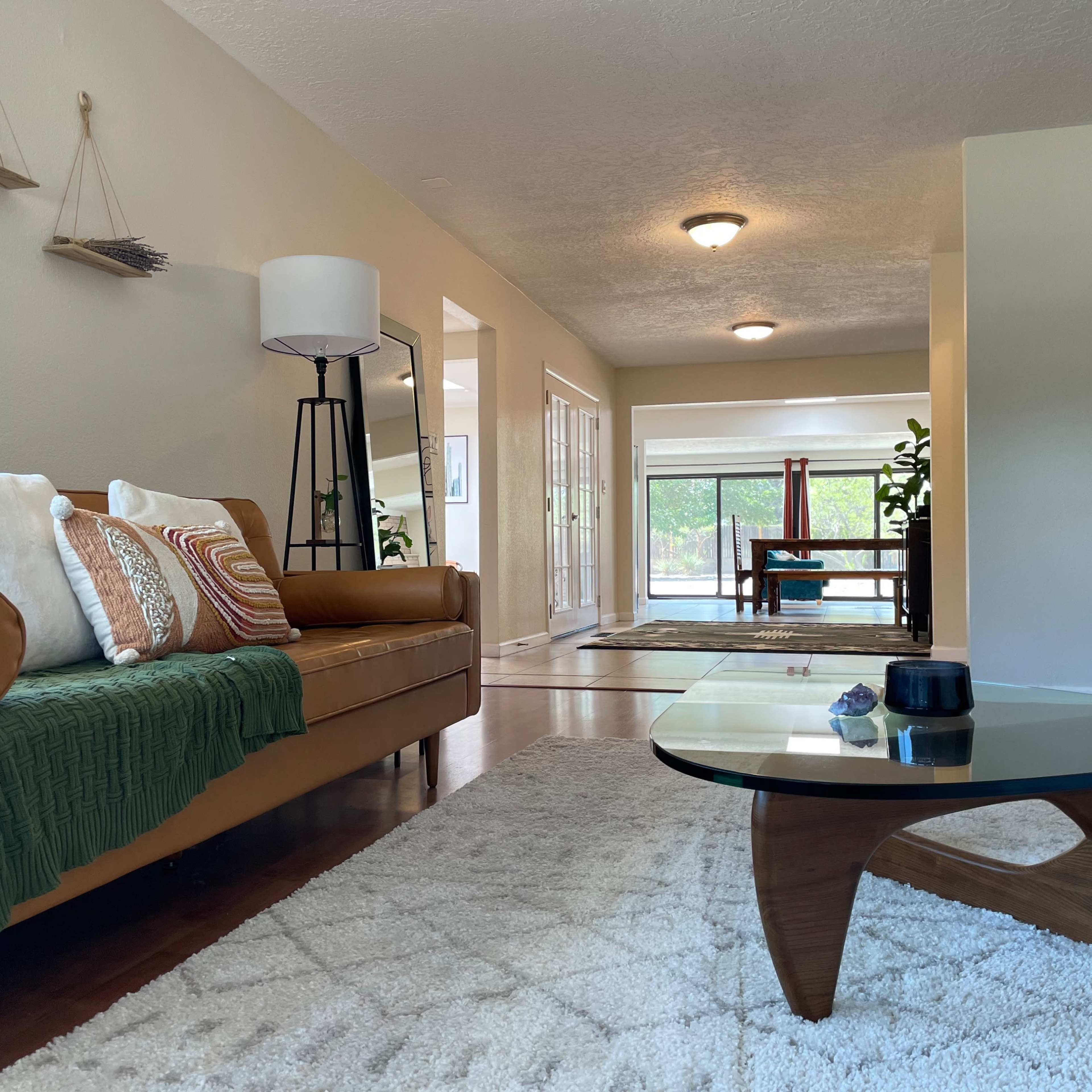 A modern living room features a tan leather sofa with a green throw, a glass coffee table, and a view of an adjacent room with a dining table.