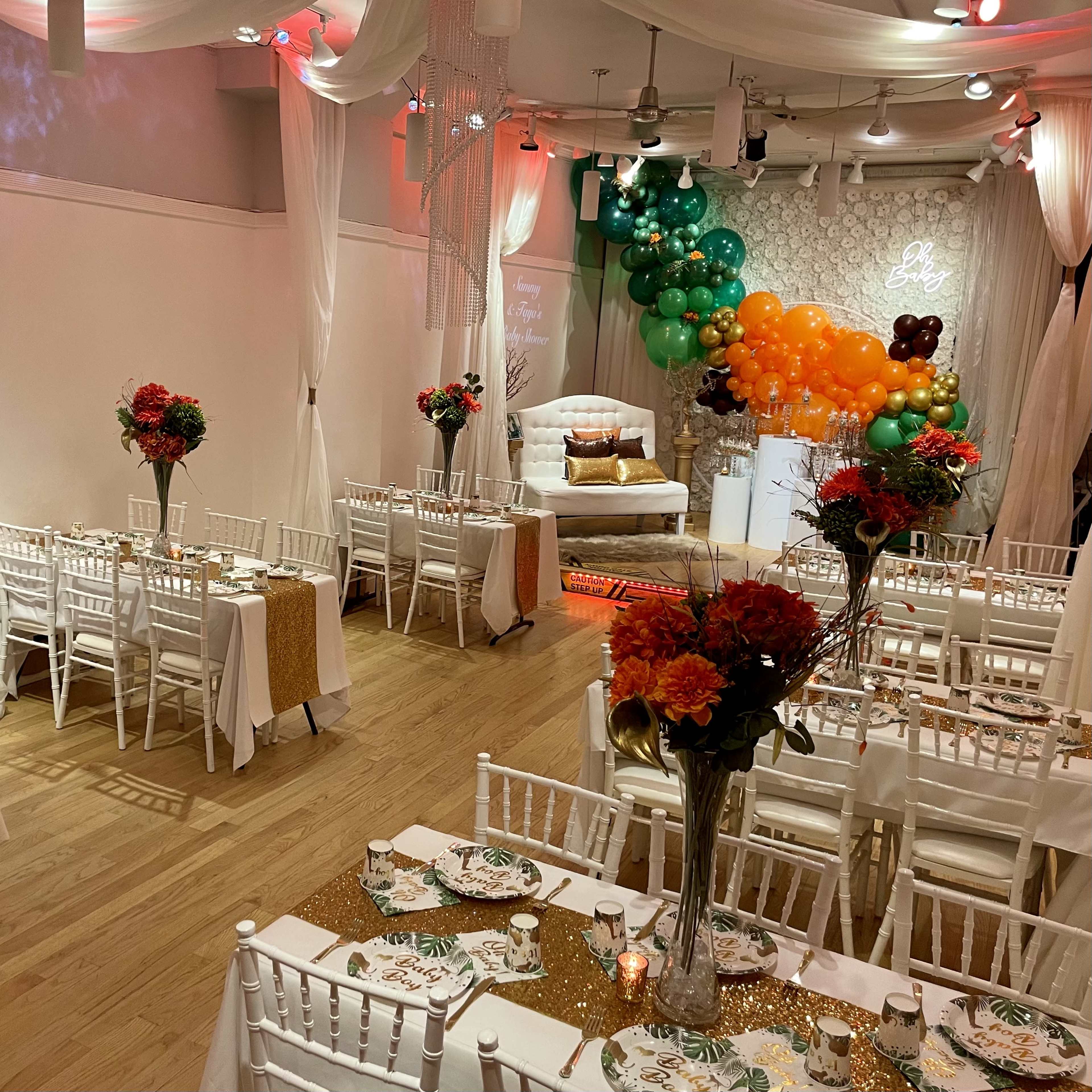 A decorated event space with white chairs arranged at tables covered in white and gold linens, a backdrop of balloons, and a seating area featuring a white sofa.