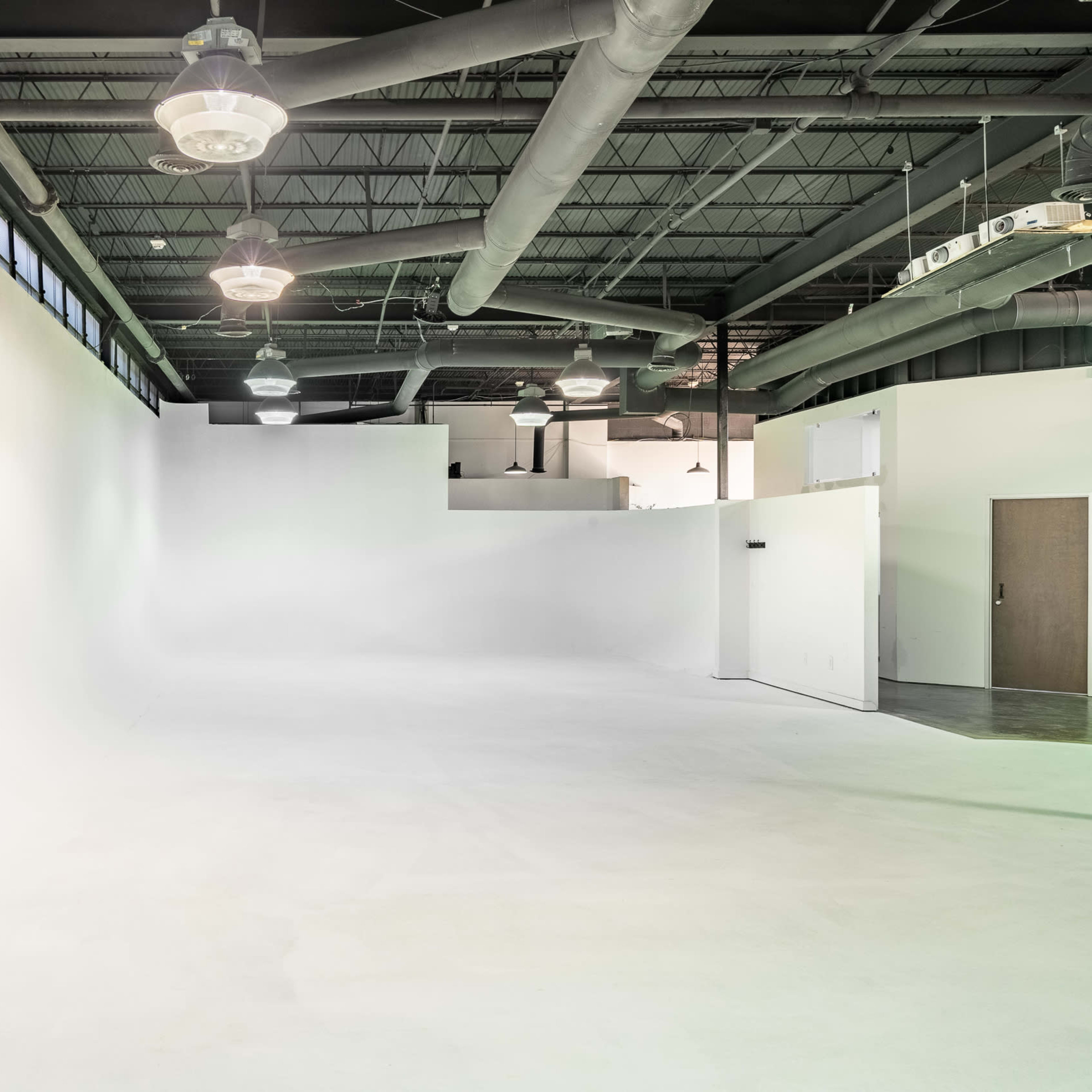 A large, empty studio space with white walls, a smooth floor, and industrial lighting fixtures.