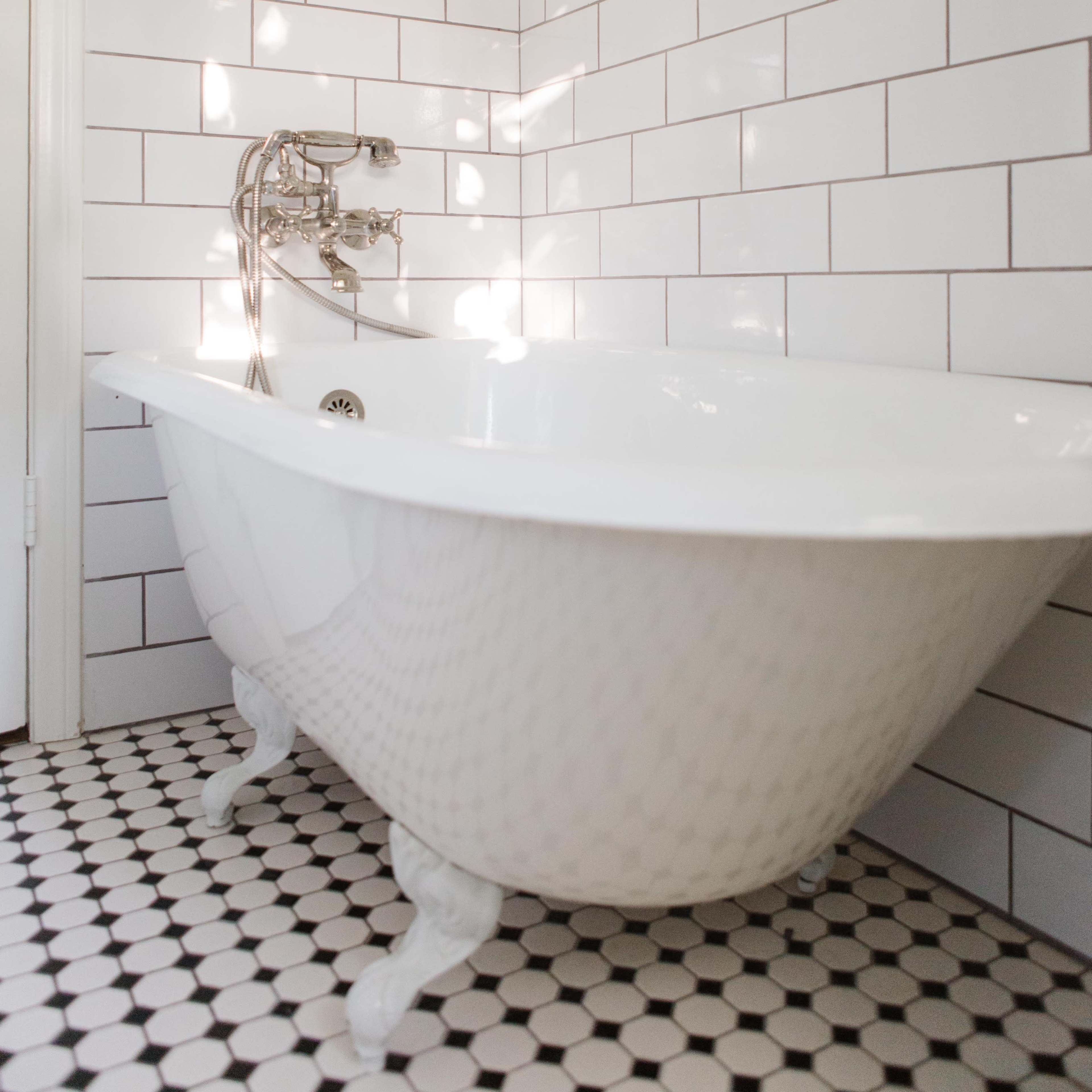 A white clawfoot bathtub positioned in a bathroom with white tiled walls and a black and white hexagonal tiled floor.