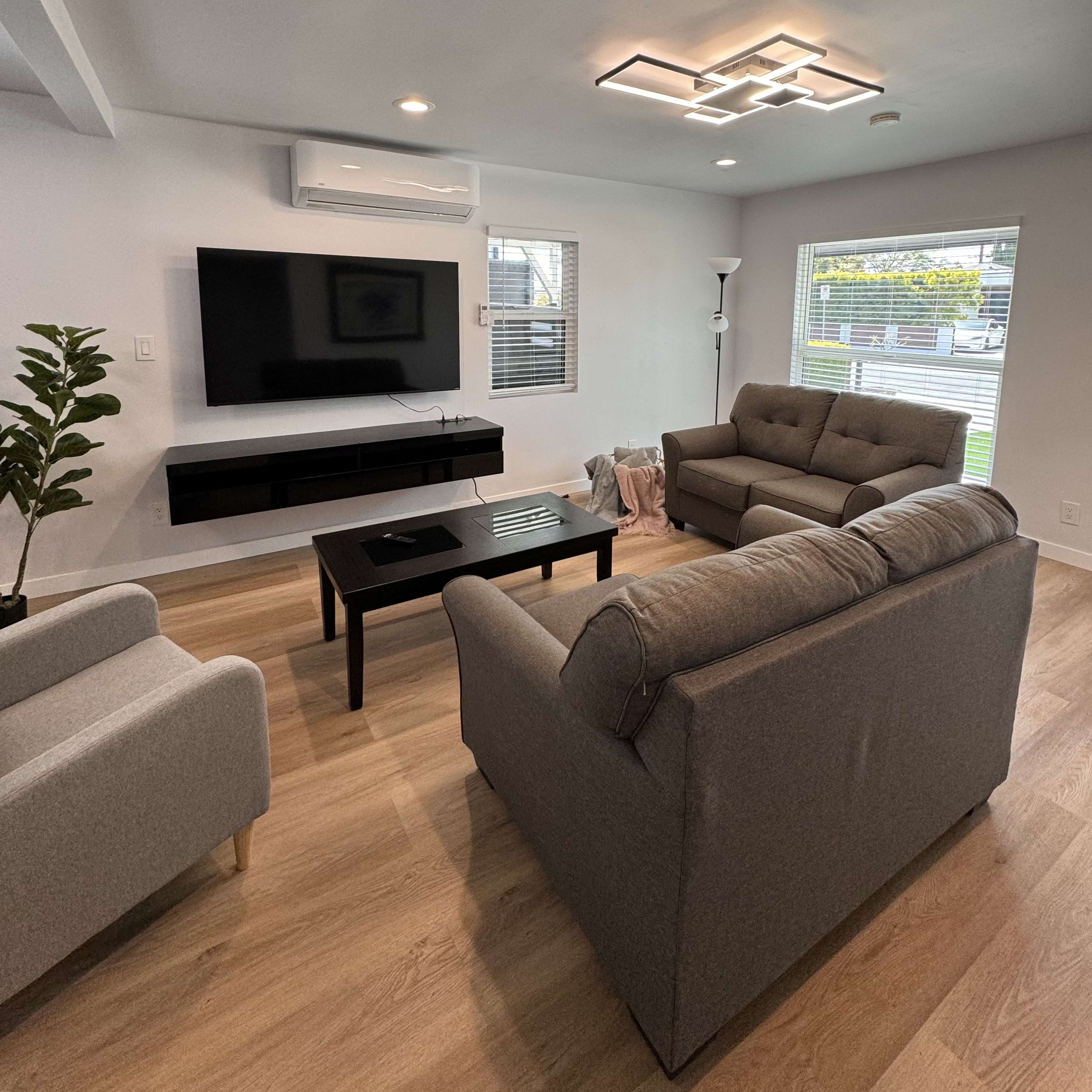 A modern living room features a gray sofa and armchair around a black coffee table, with a wall-mounted television and an air conditioning unit.