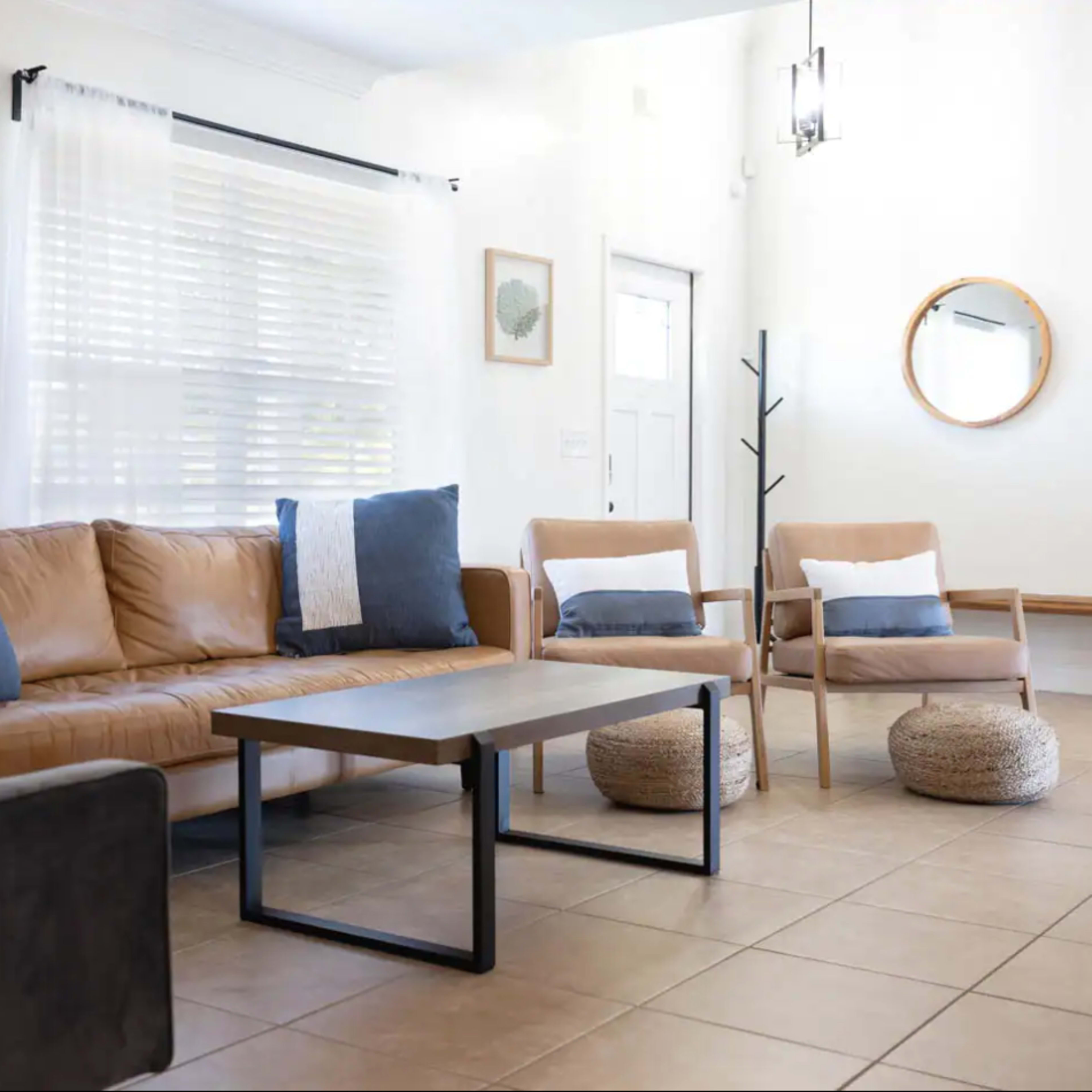 A living room features a brown leather sofa, two armchairs, a round mirror, and a coffee table on tiled flooring.