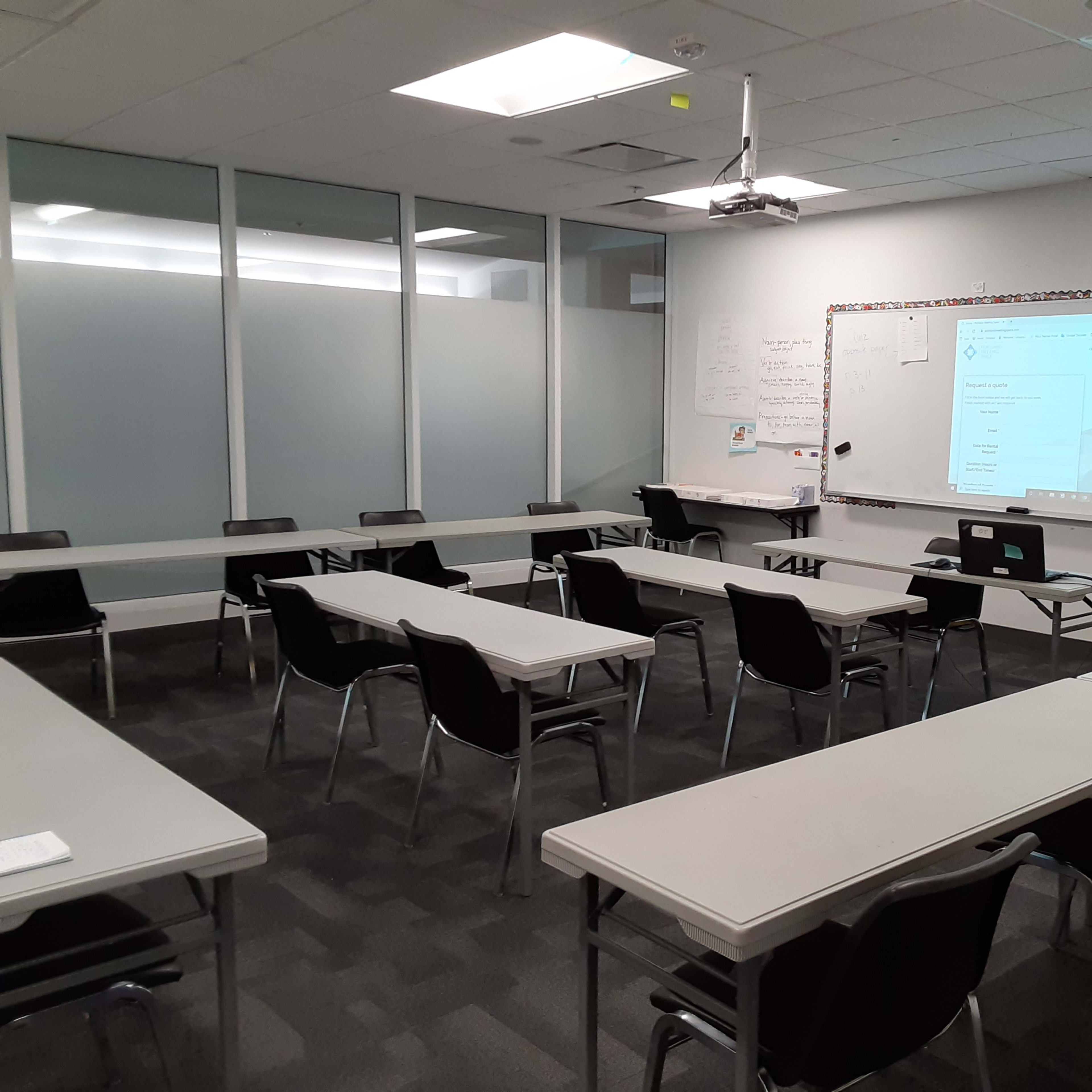 A classroom with several white tables arranged in rows, black chairs at each table, and a whiteboard displaying a presentation on one wall.