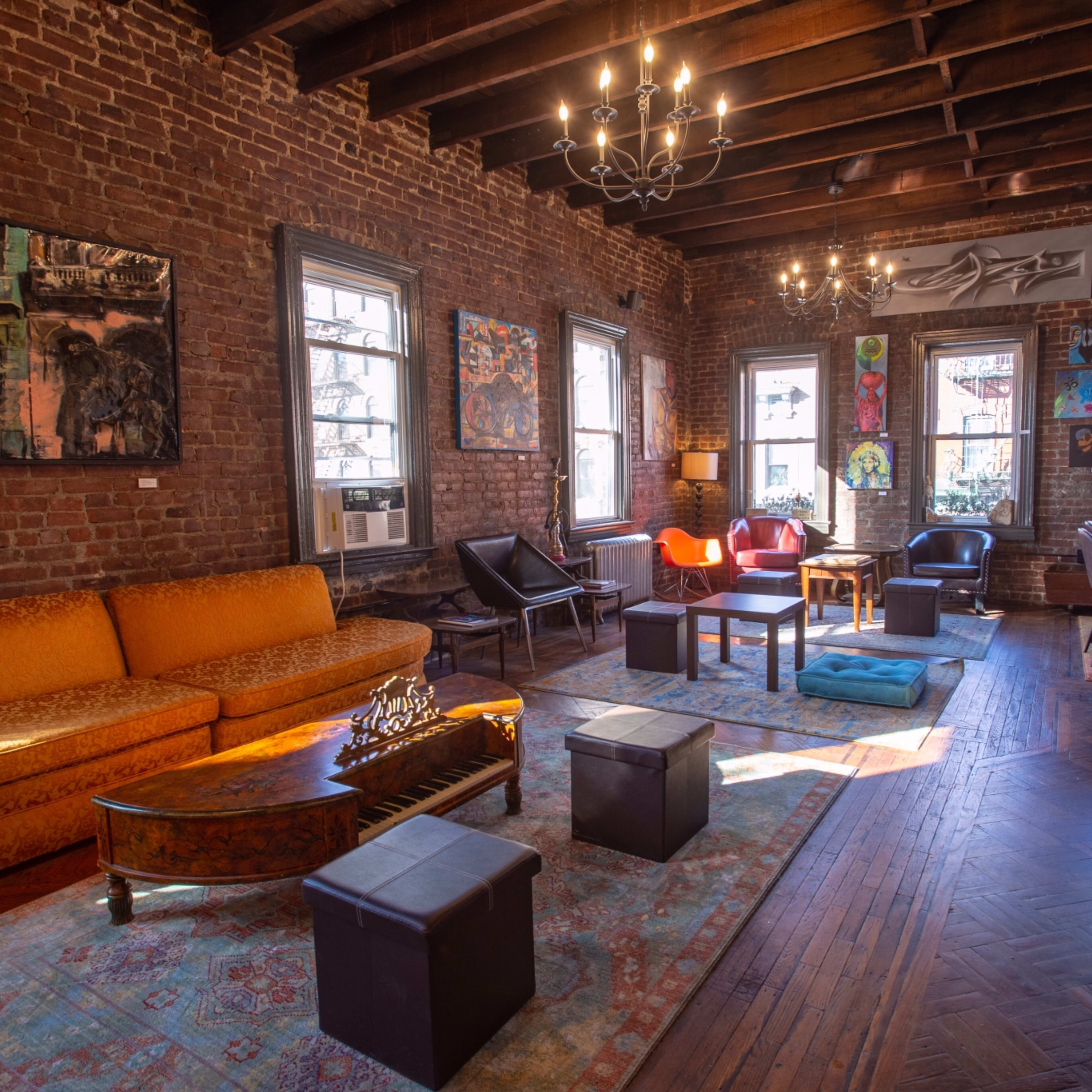 The room features exposed brick walls, a large orange sofa, various artworks, and a mix of seating arrangements, illuminated by overhead lighting.