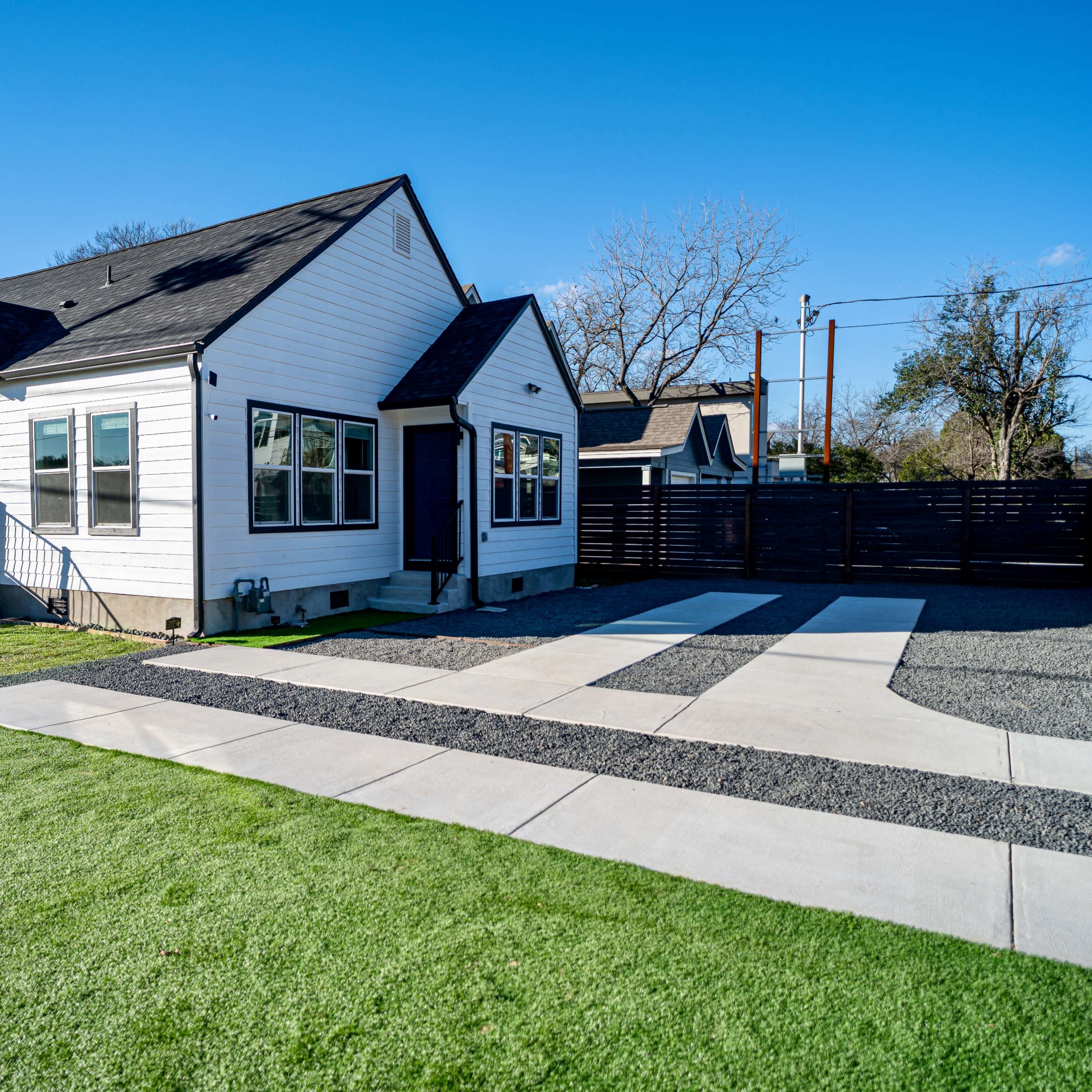 A white house with multiple windows and a blue door sits beside a gravel driveway, surrounded by green grass and a wooden fence.