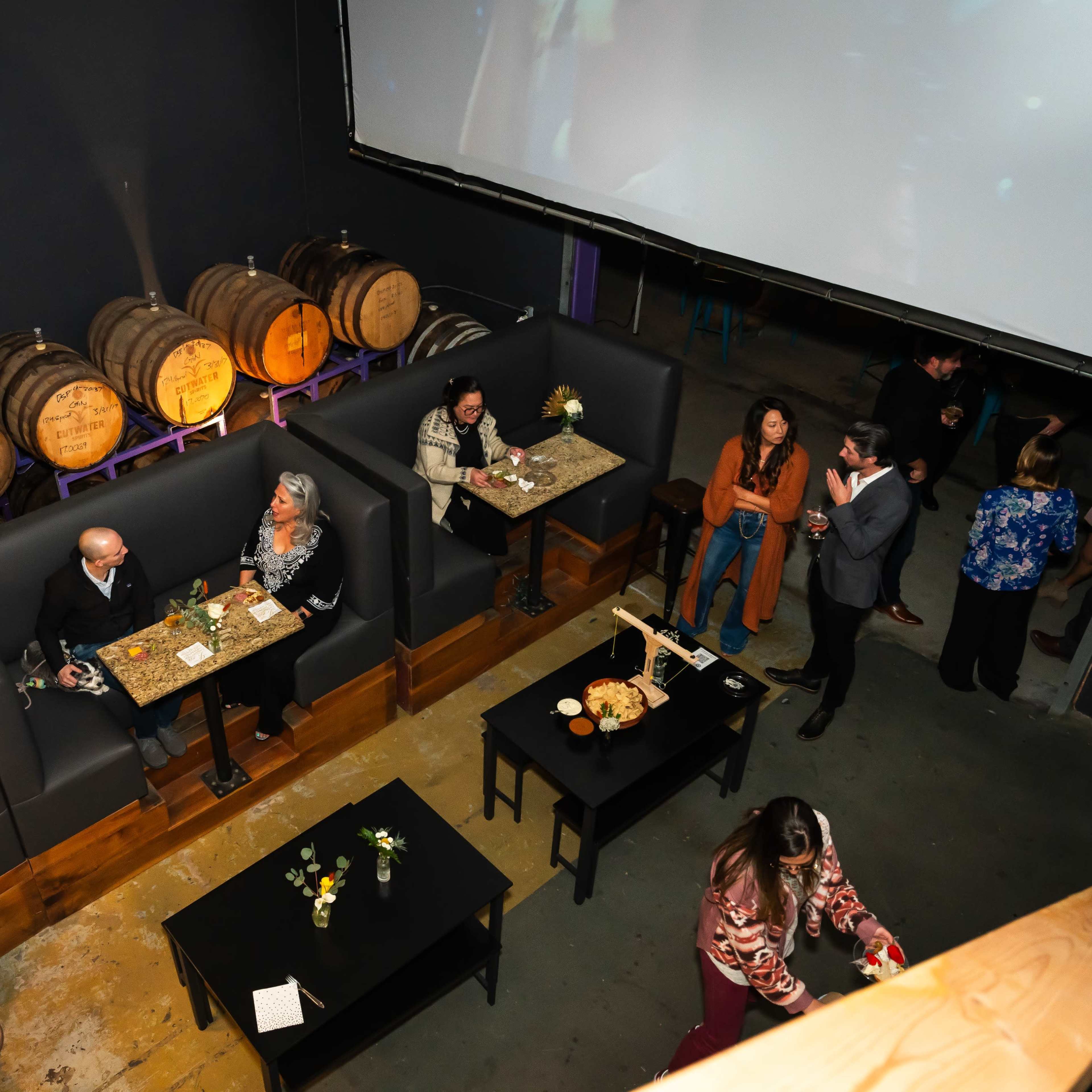A dimly lit bar setting with patrons seated at tables, surrounded by wooden barrels and a large screen displaying a video.