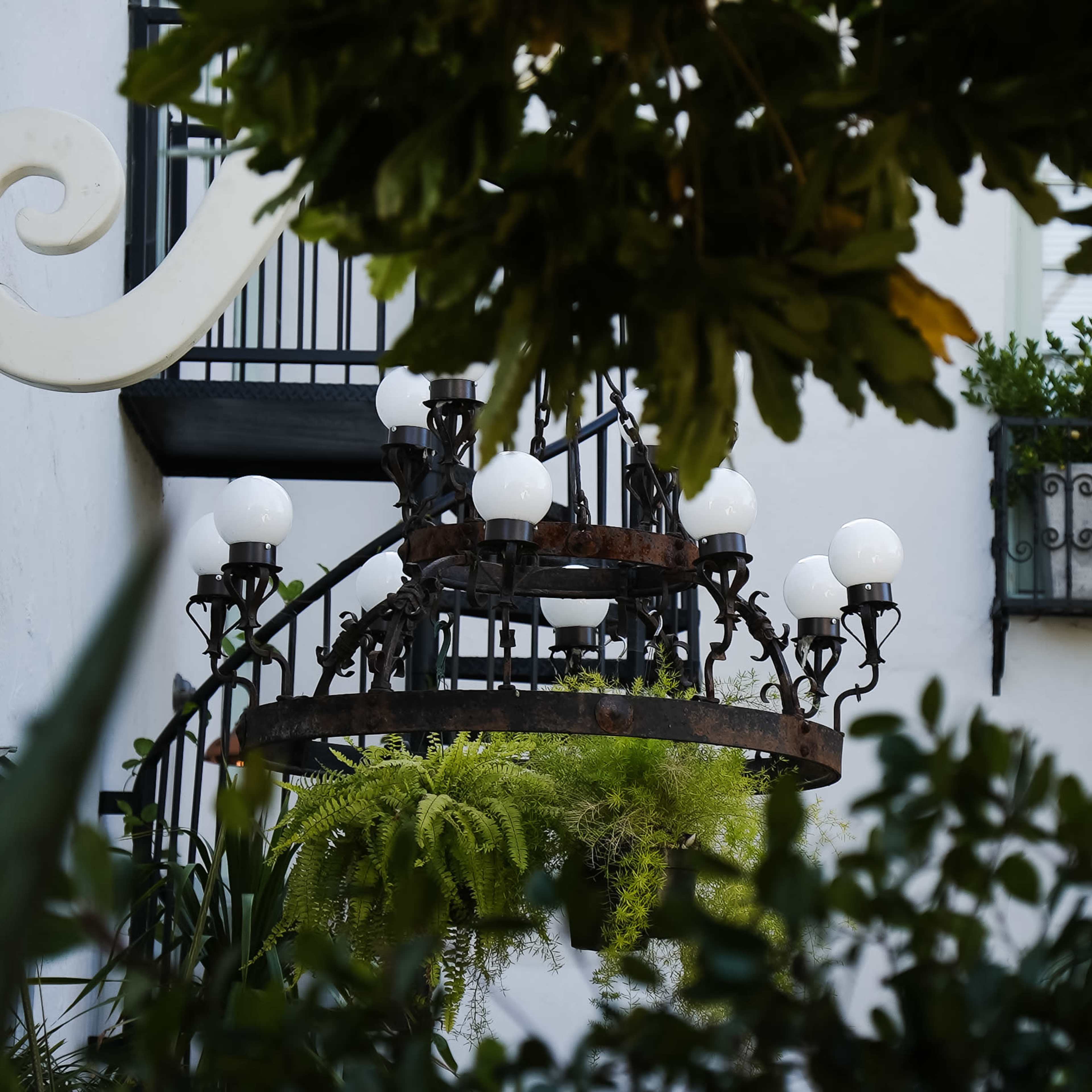 A wrought iron chandelier with white globes hangs in a verdant courtyard near a balcony with potted plants.