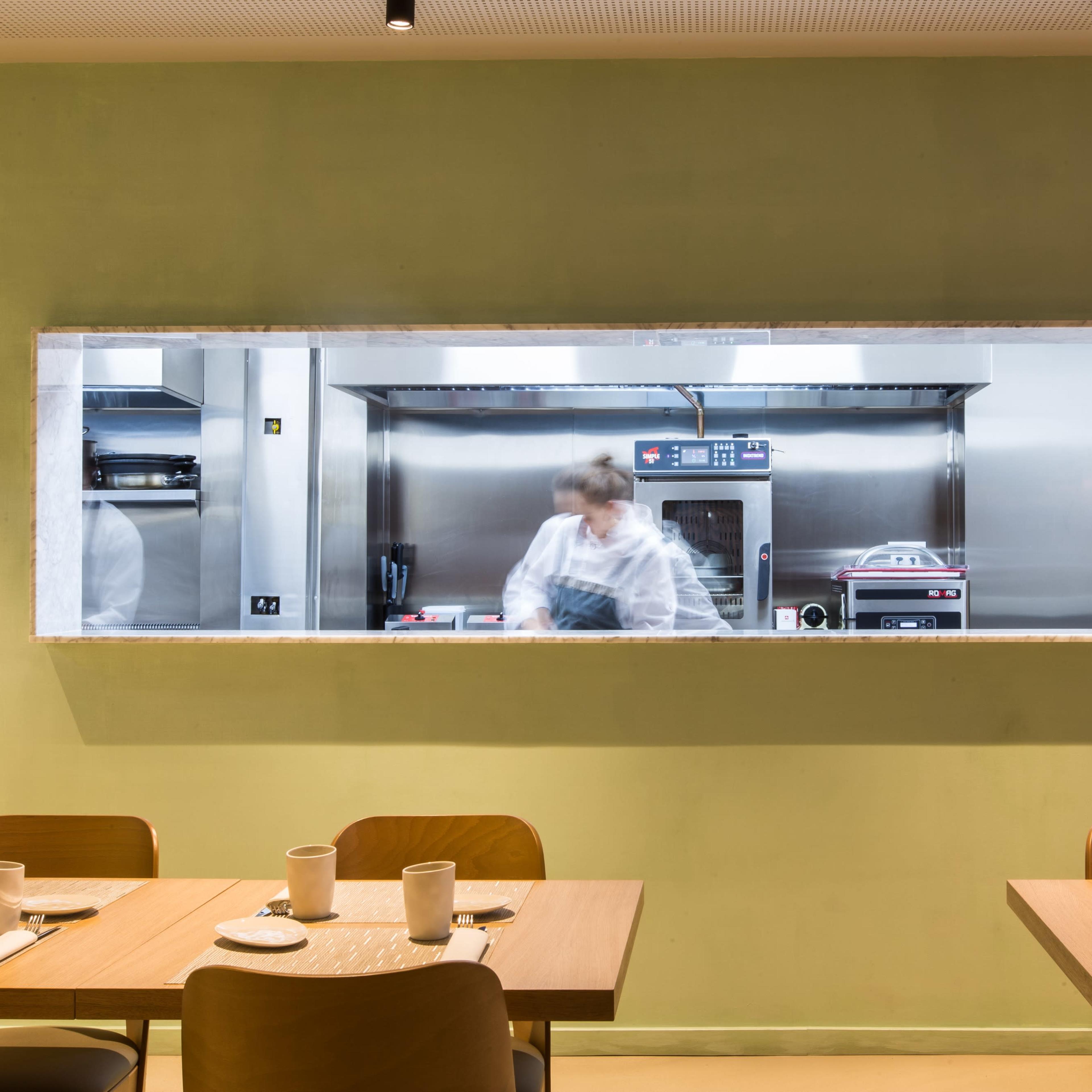 A chef is working in a modern kitchen visible through a rectangular window, with wooden tables set for diners in the foreground.