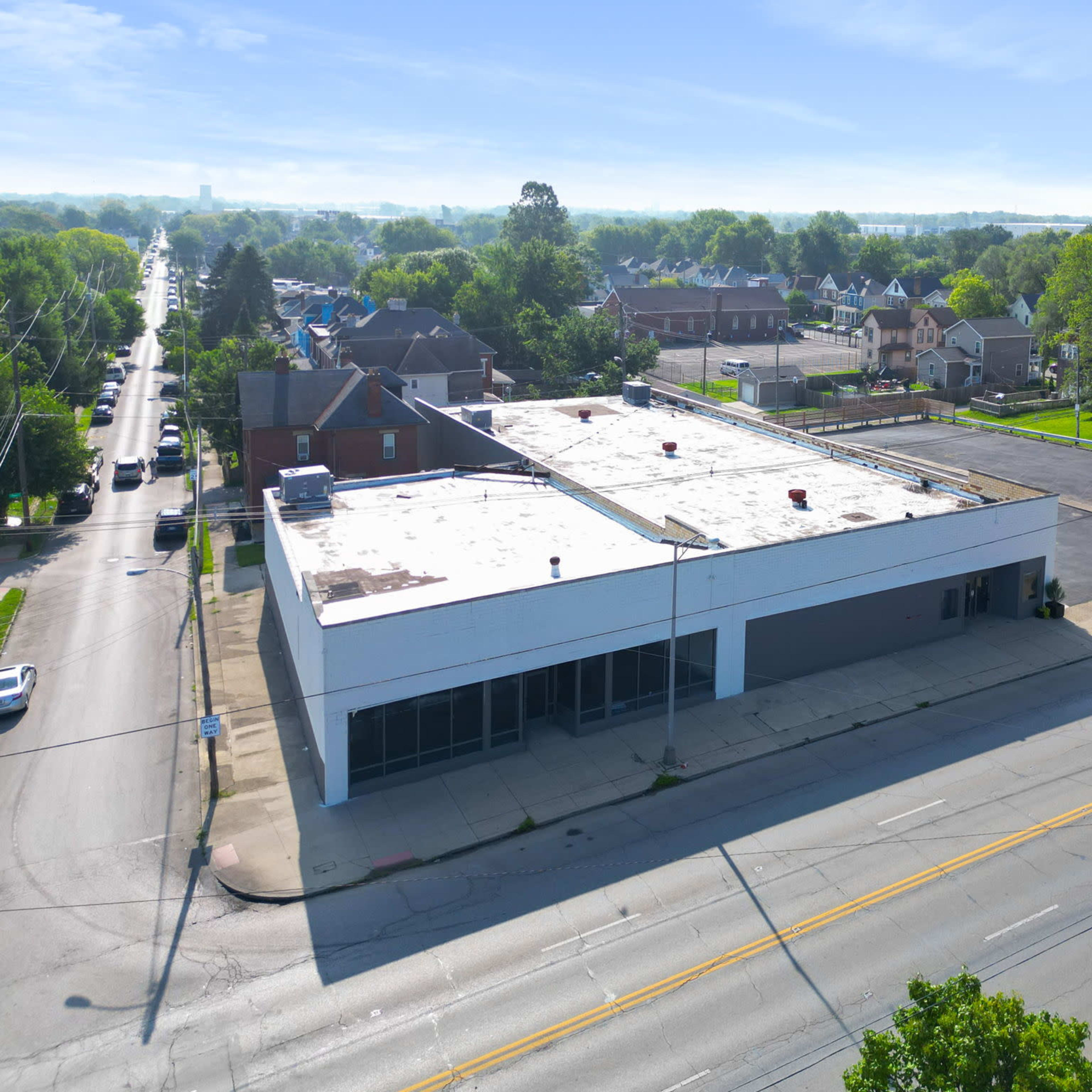 An aerial view shows a vacant commercial building at the corner of a street, surrounded by residential homes and a parking lot.