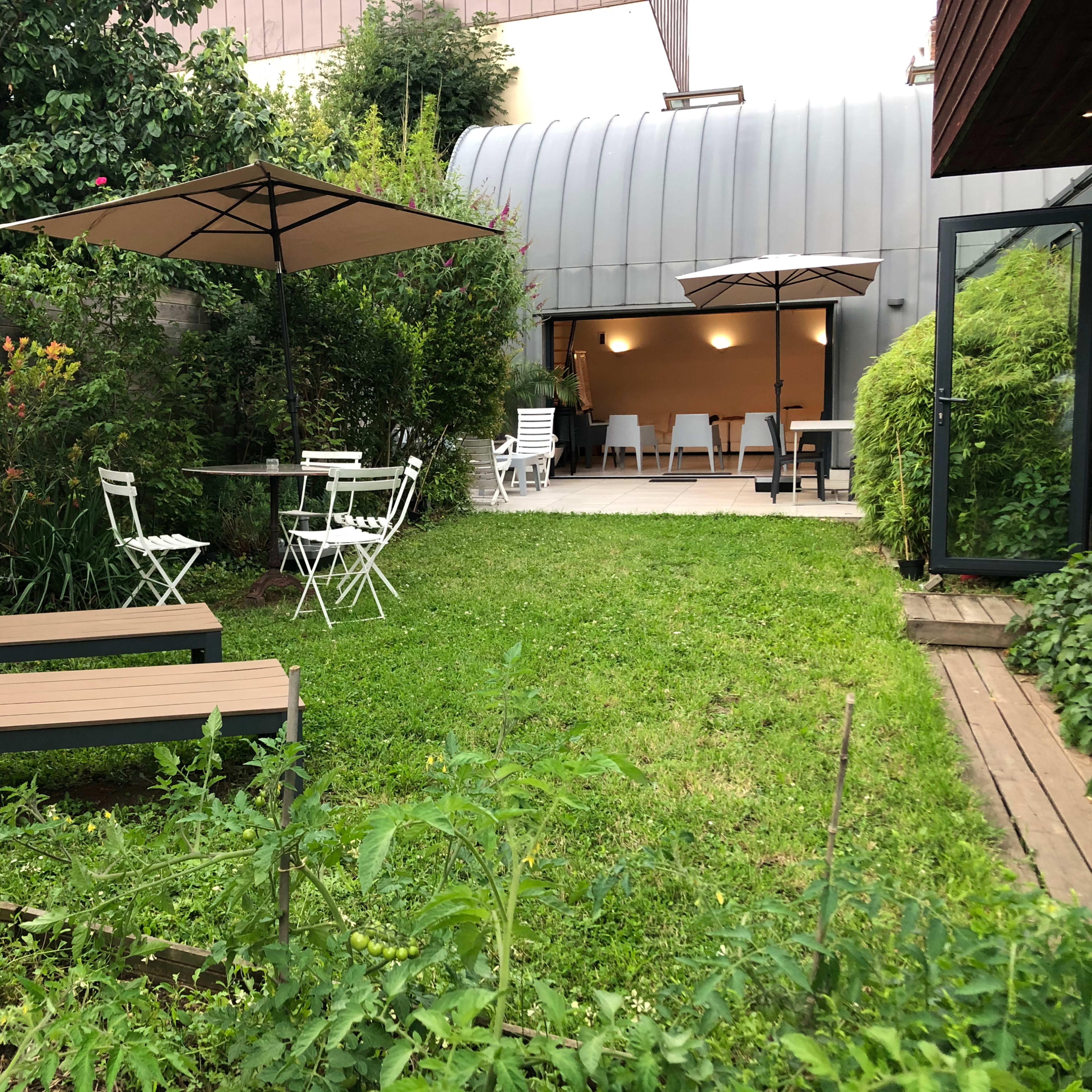A well-maintained garden features green grass, two lounge chairs, a dining table with chairs, and two umbrellas, adjacent to a modern building.