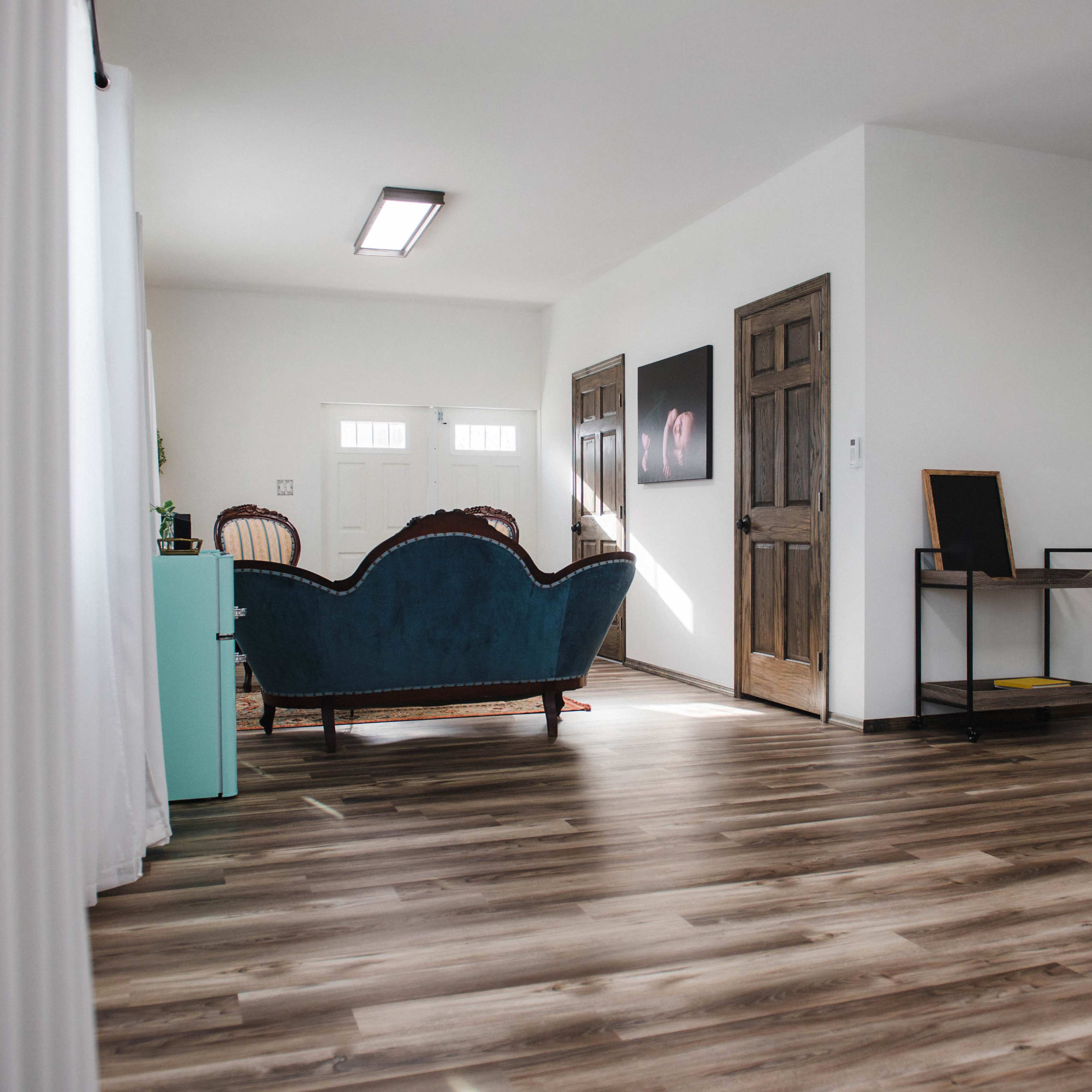 A blue couch is positioned in a minimalist room with wooden floors and doors leading to an entryway.