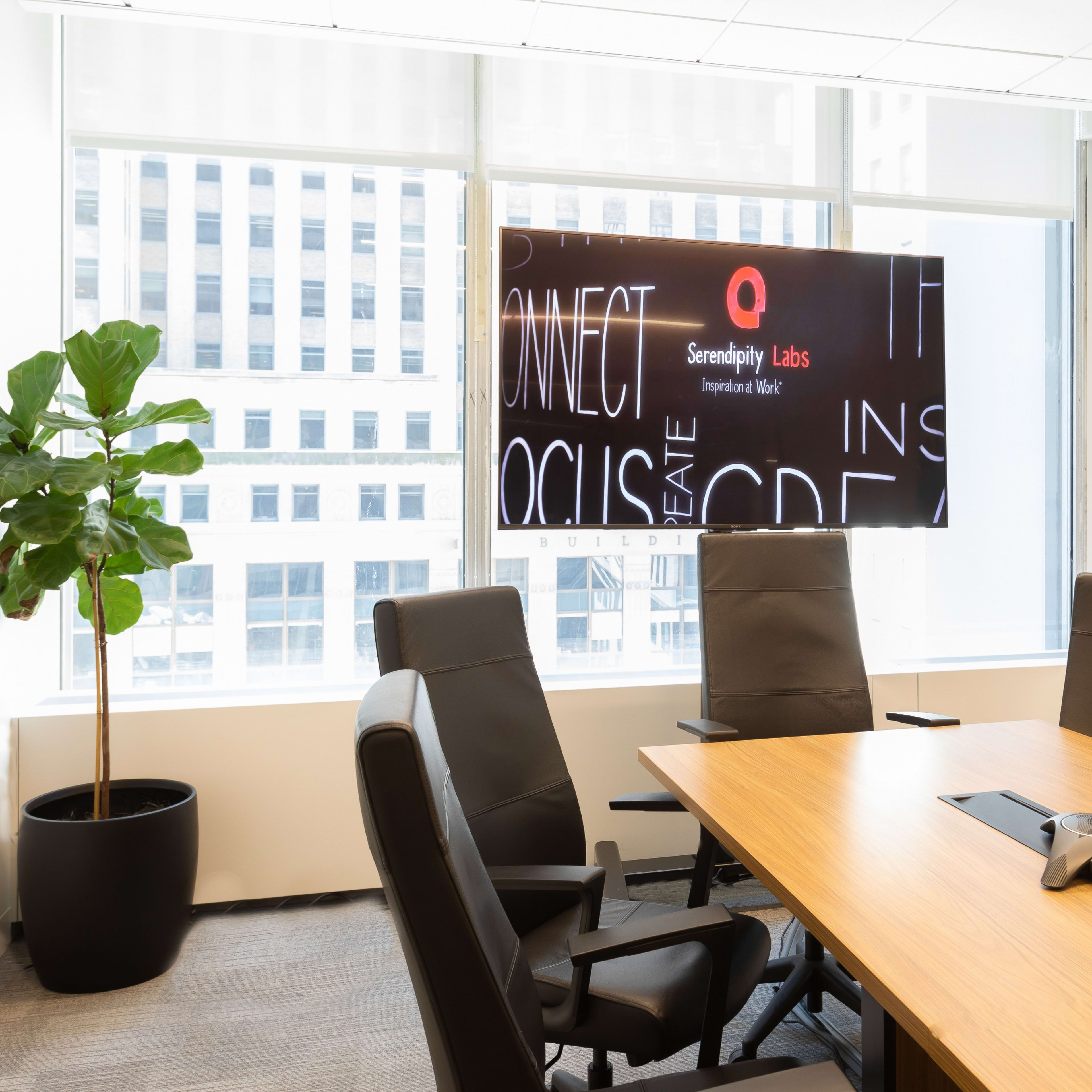 A modern conference room features a large wooden table, black office chairs, a speakerphone, and a wall-mounted screen displaying graphics, alongside a potted plant.