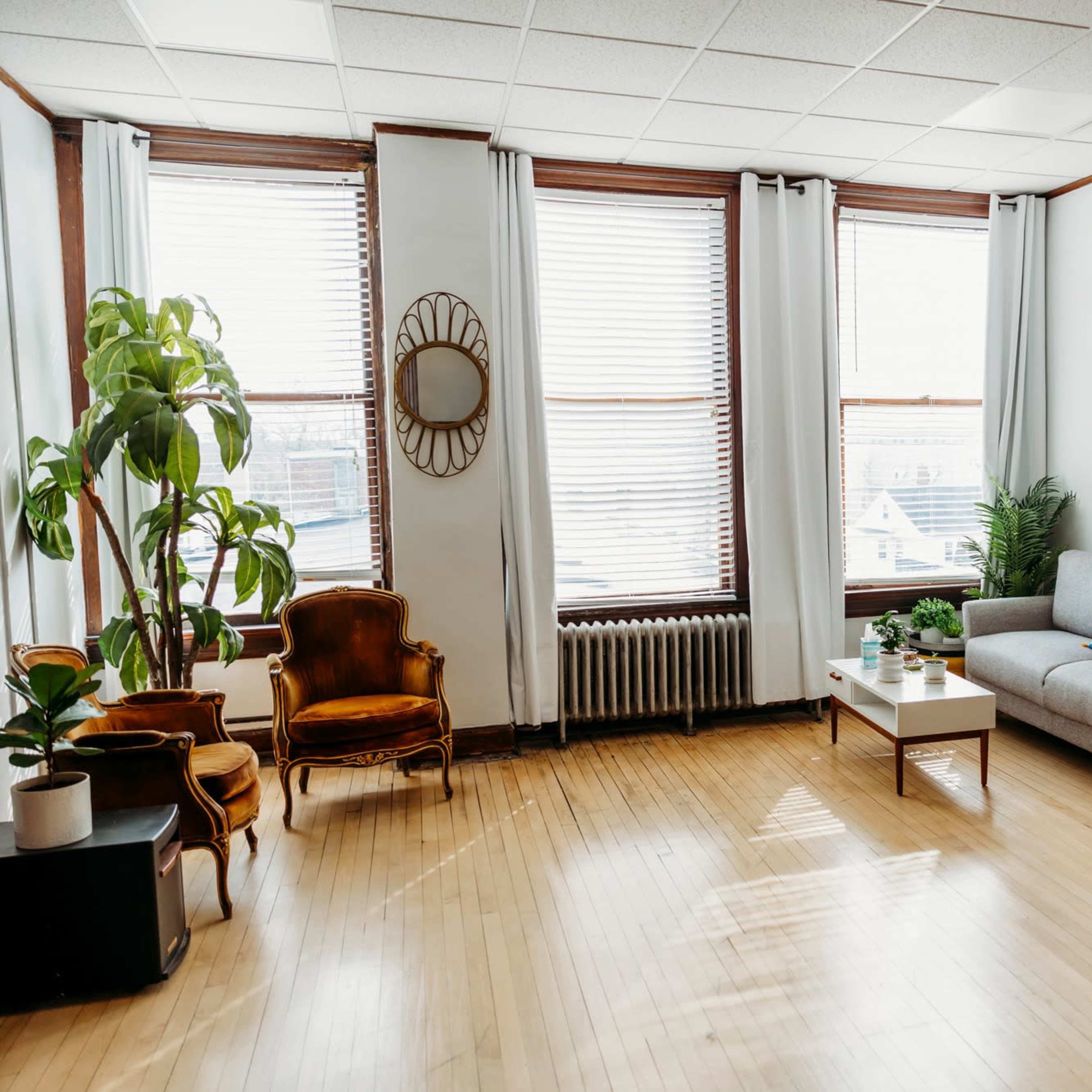 A bright living room features large windows, a gray sofa, wooden furniture, and indoor plants.