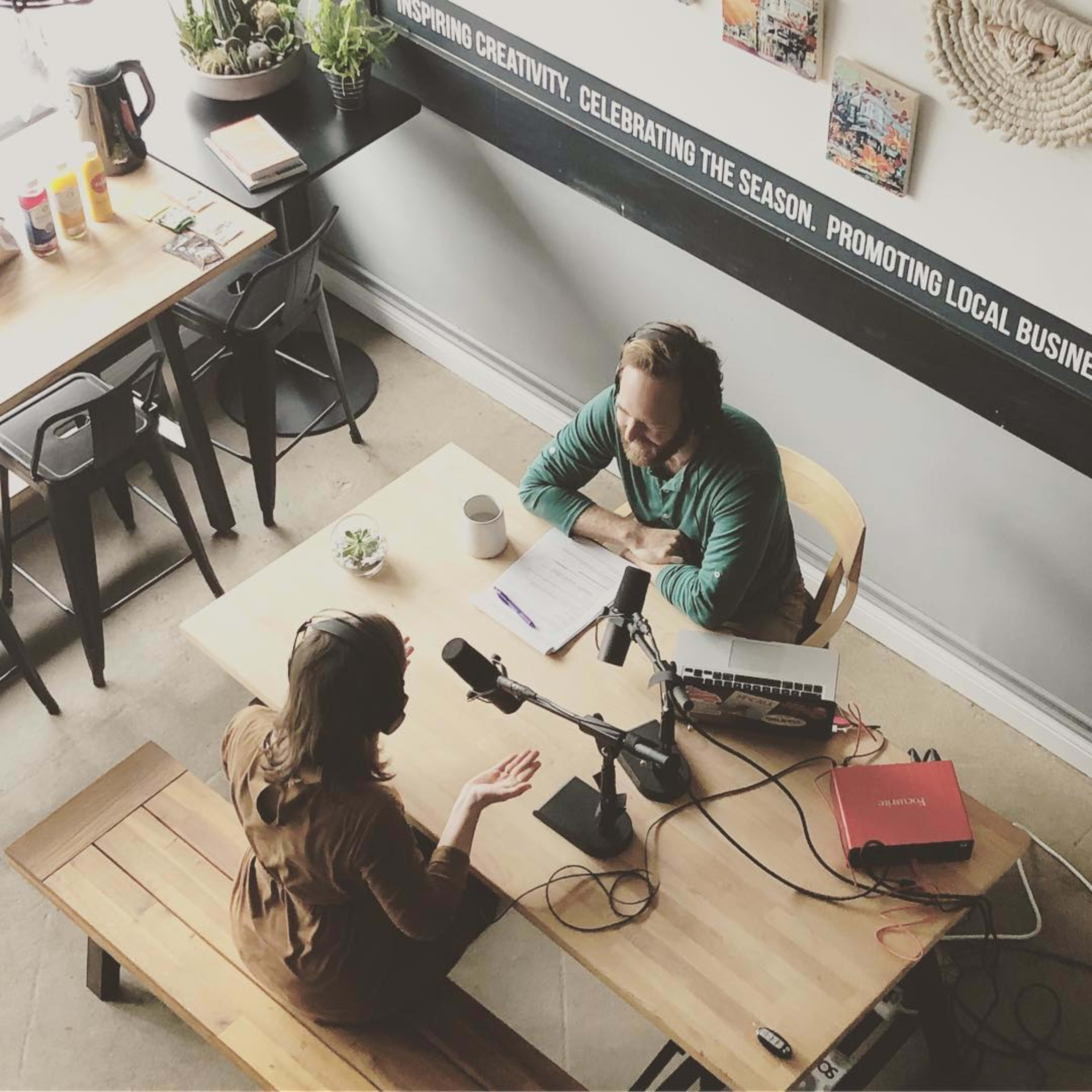 Two people are engaged in a conversation at a wooden table in a casual setting, with a microphone and notes in front of them.