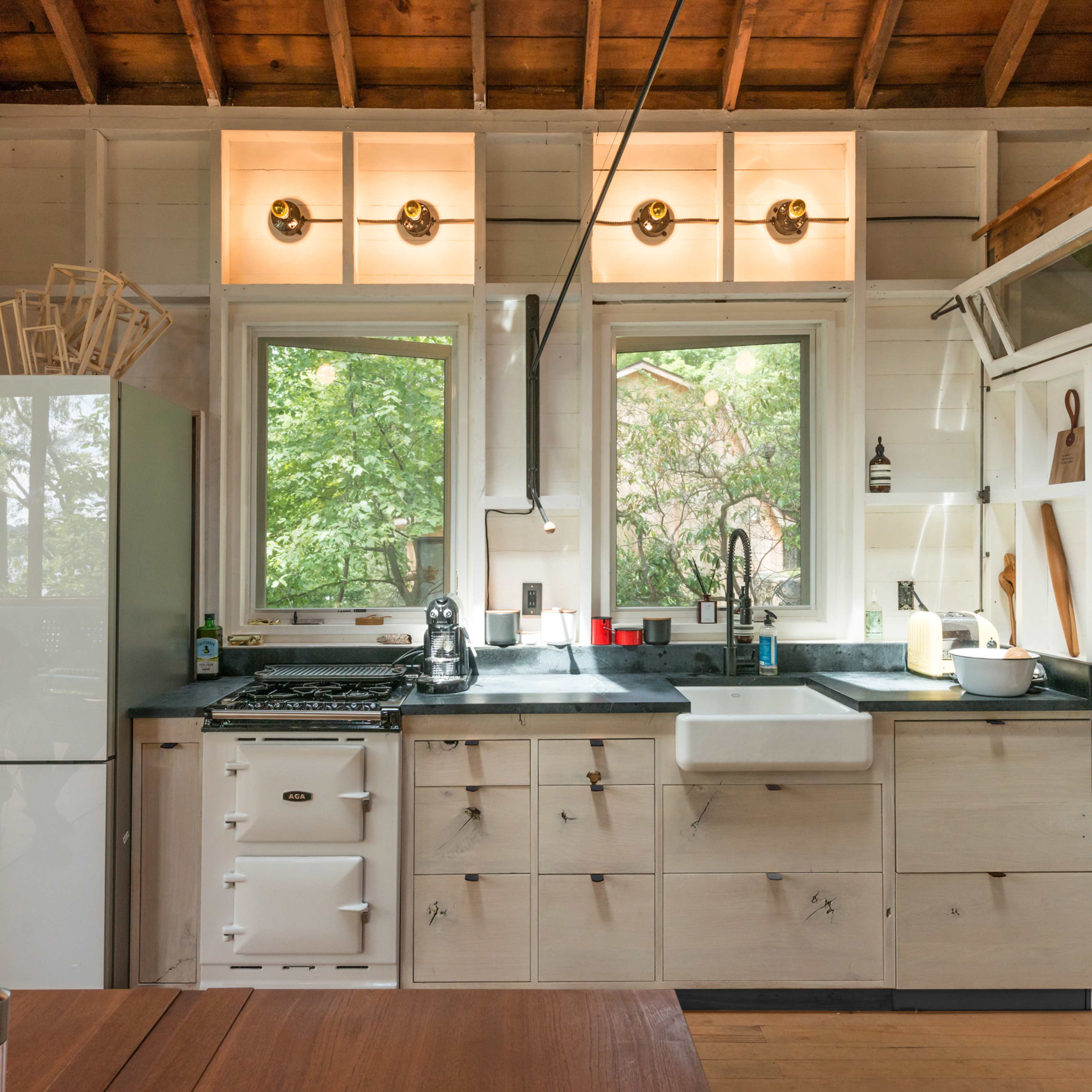 A well-lit kitchen featuring a white refrigerator, a sink, a gas stove, and wooden cabinetry against a backdrop of large windows.