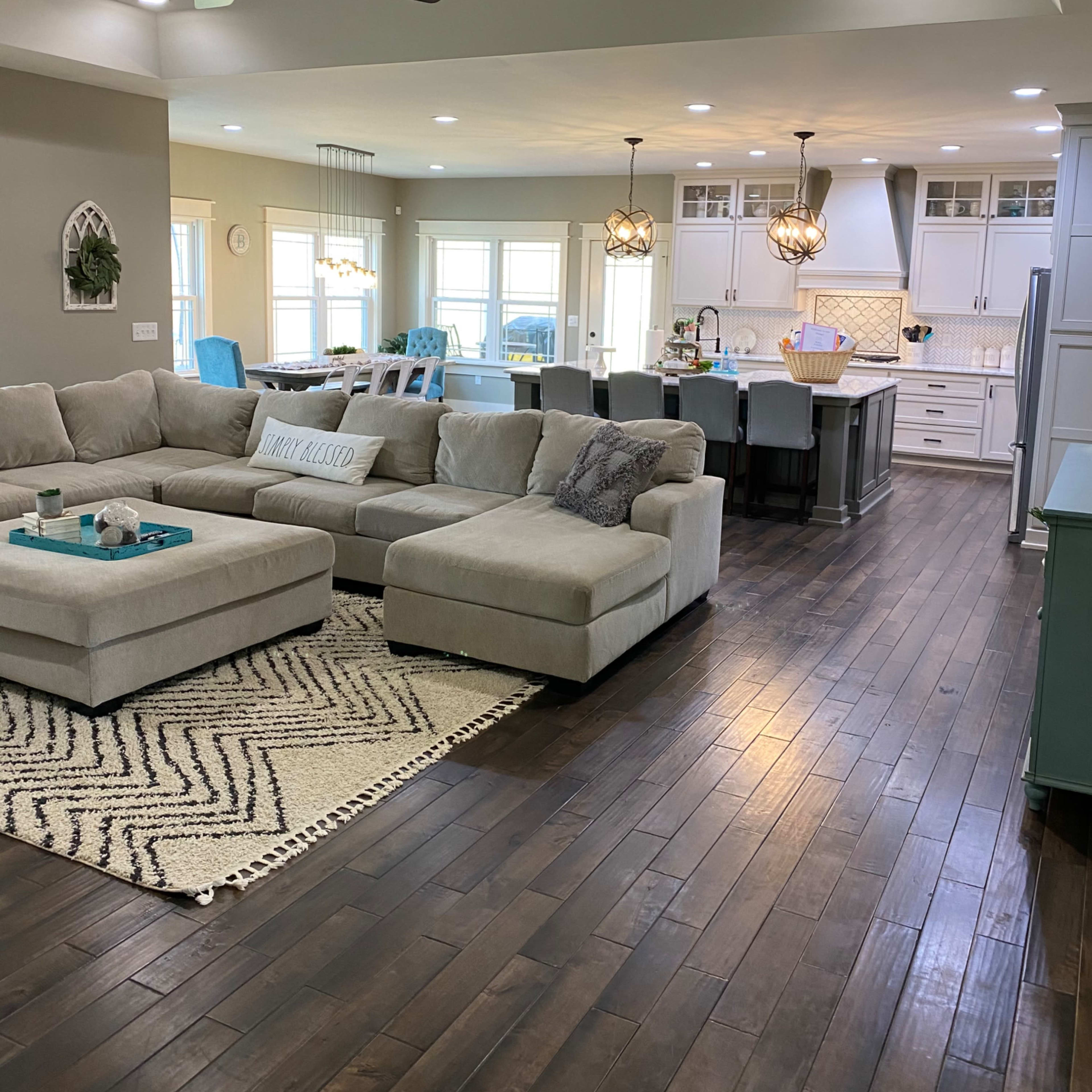 A modern living room with a large sectional sofa, a decorative rug, and an open kitchen in the background featuring a central island and pendant lighting.