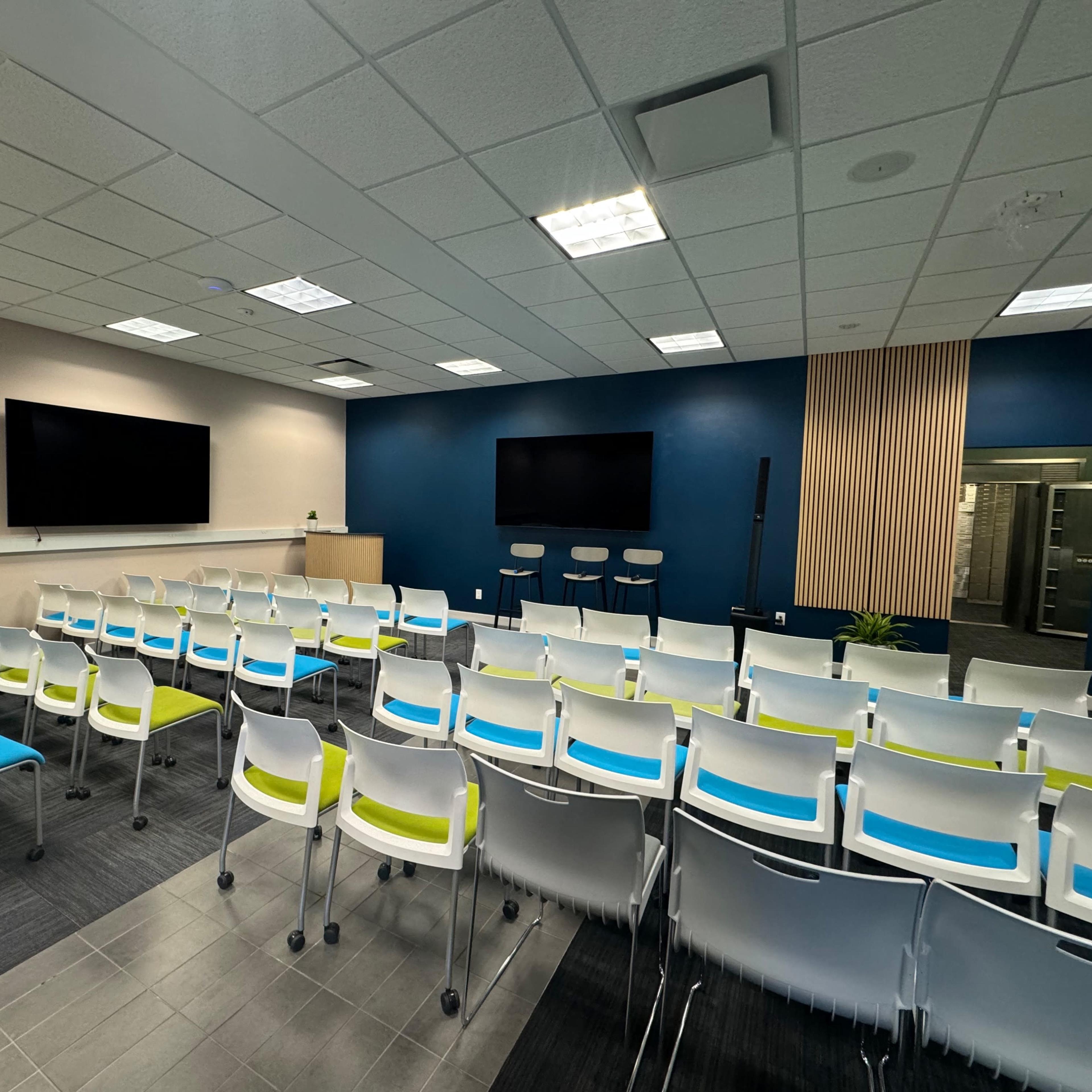 A modern conference room with rows of white chairs featuring blue and green accents, two large screens on the wall, and a backdrop of blue paint and wooden paneling.