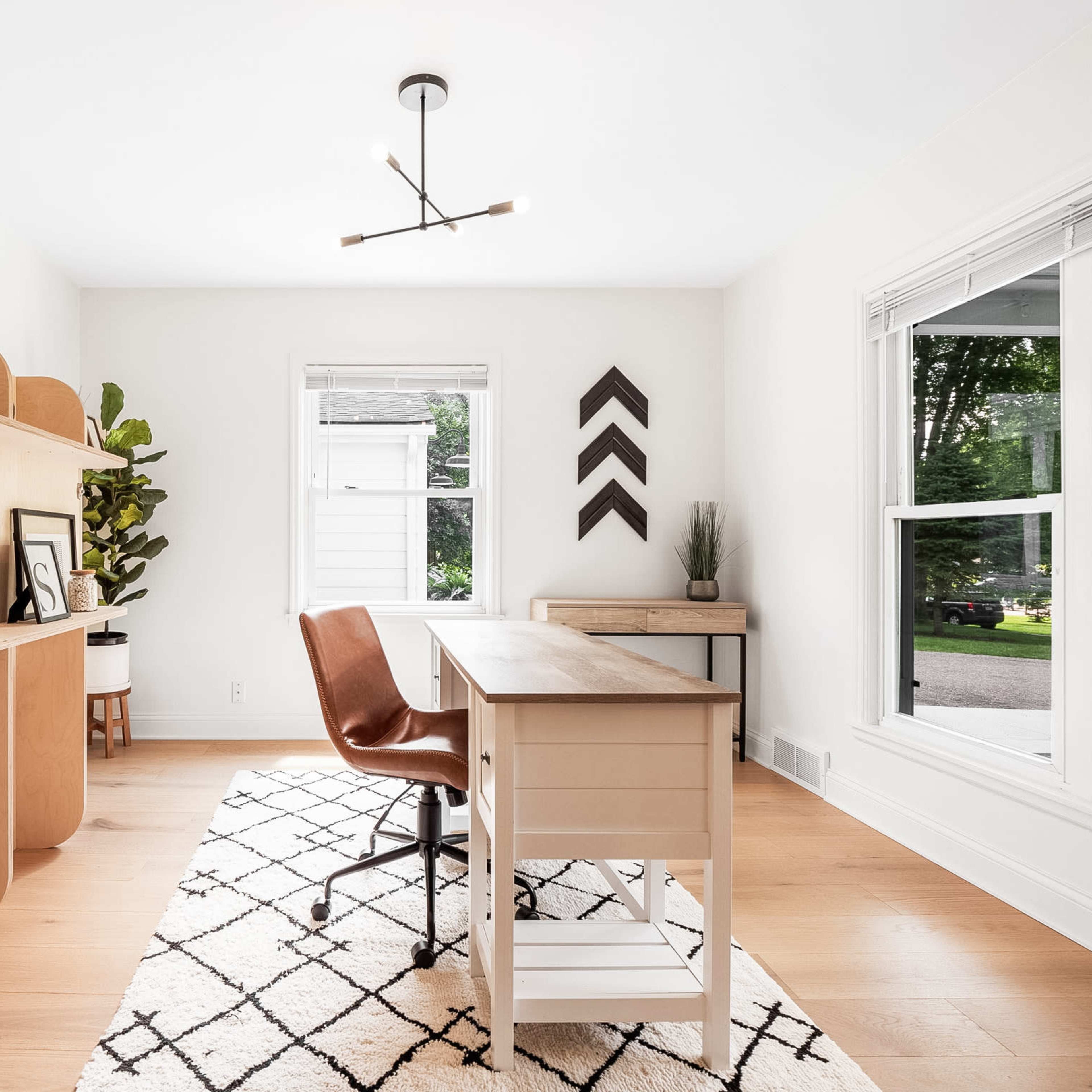 A modern home office featuring a wooden desk with a leather chair, a large shelving unit, a potted plant, and large windows allowing natural light.
