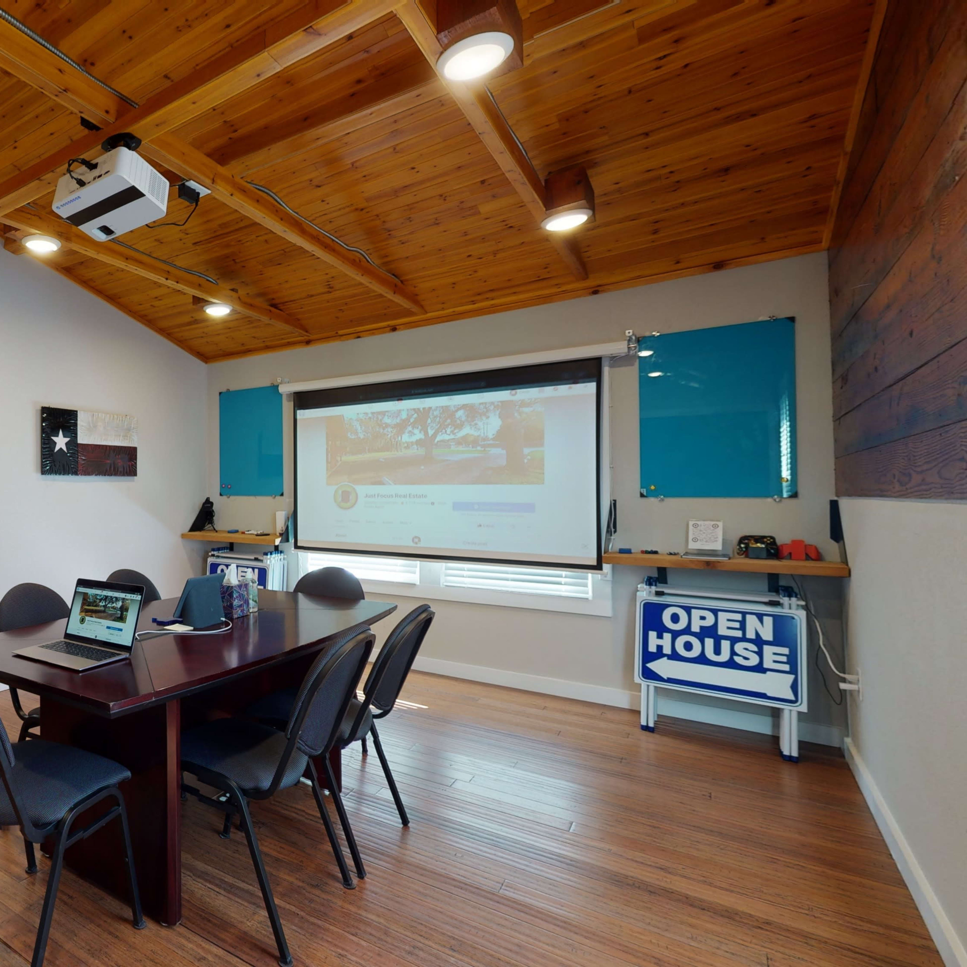 A meeting room with a wooden ceiling, a table with chairs, and a projector displaying content on a wall, leading to a living area with a couch.