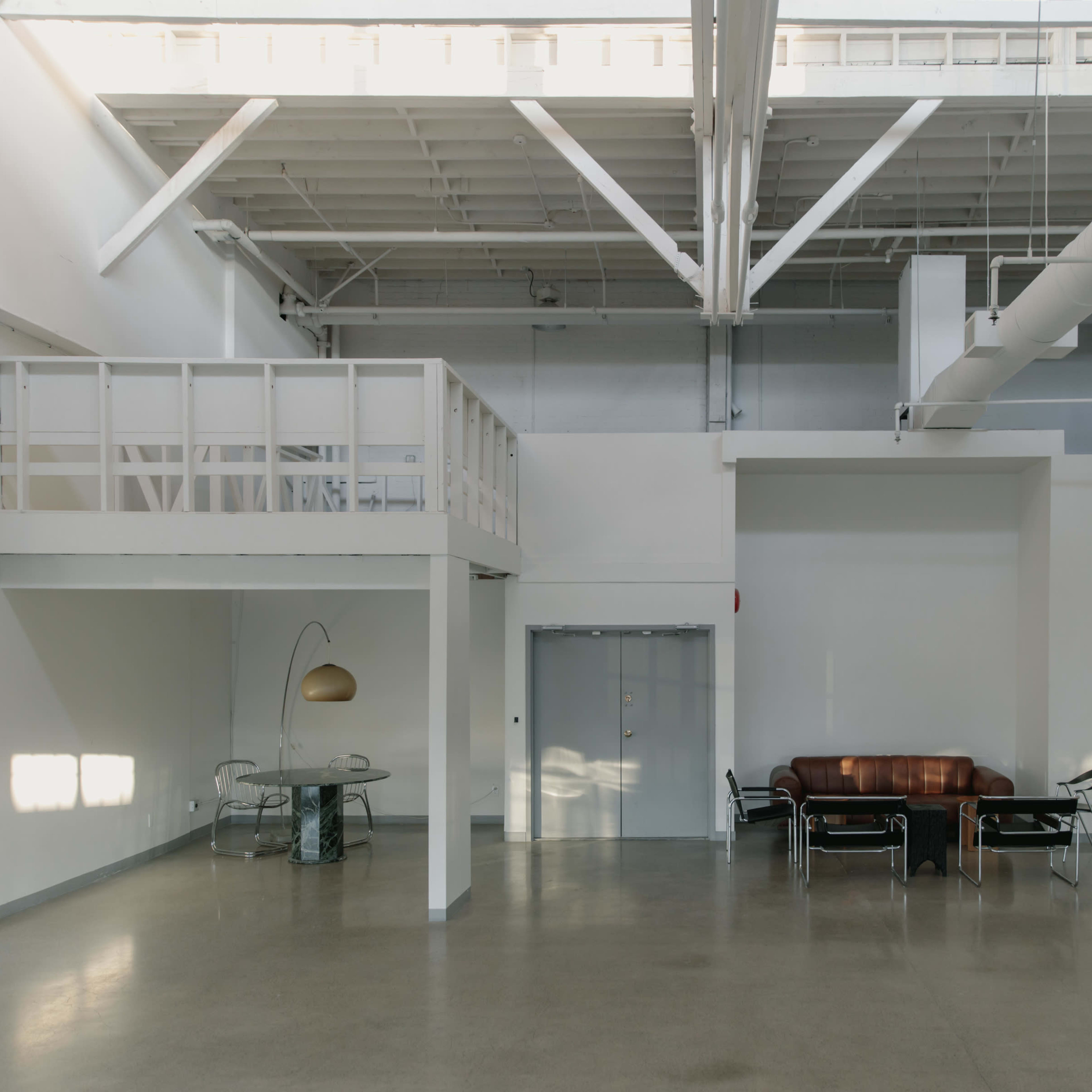 A spacious, modern interior with high ceilings, a lofted area, a brown leather sofa, a glass-topped table, and white walls.