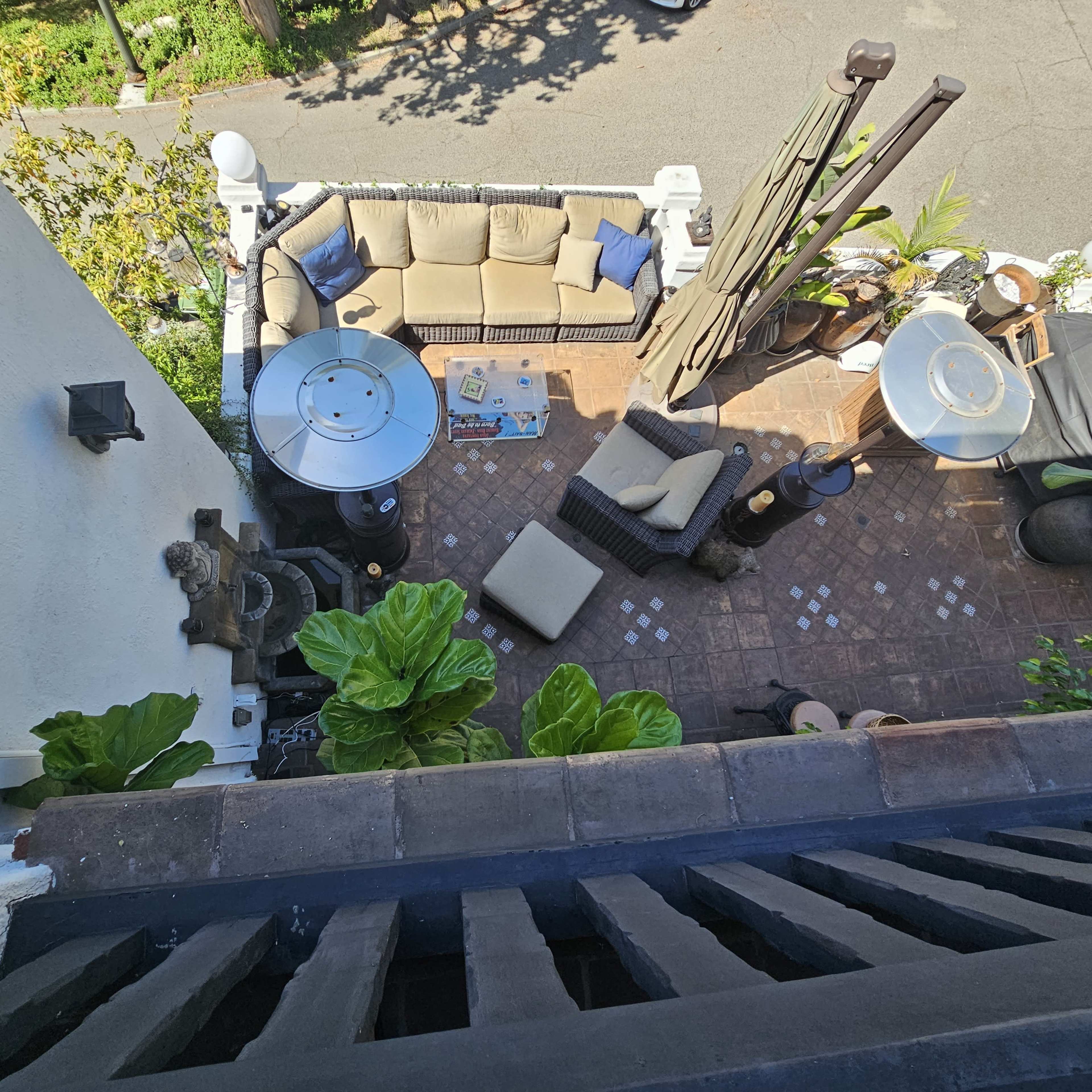 A patio area with outdoor furniture, including a couch and a table, surrounded by plants and a grill, viewed from an elevated angle.