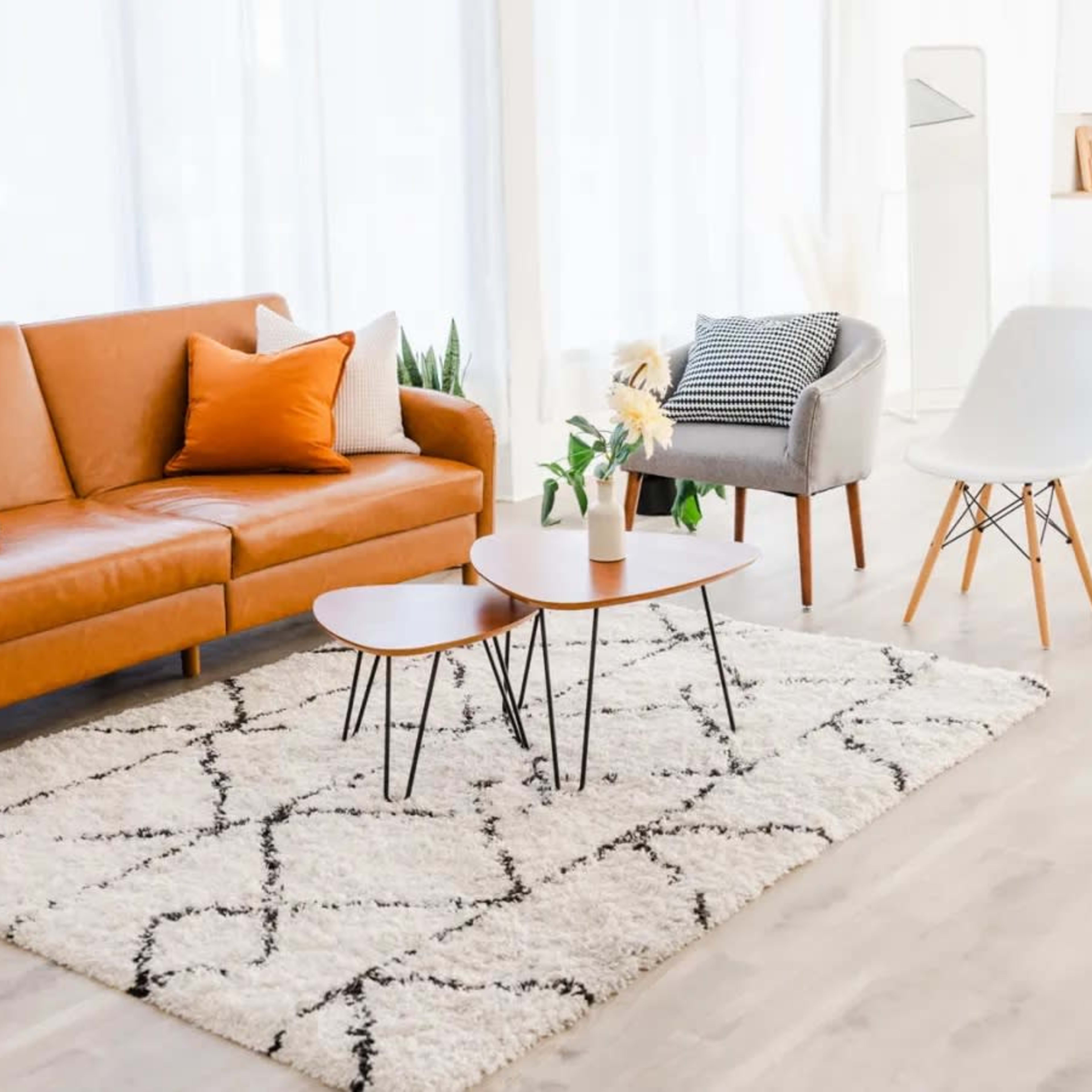 A living room with a brown leather sofa, a gray chair, a white chair, and a coffee table on a patterned rug, with light streaming through large windows.