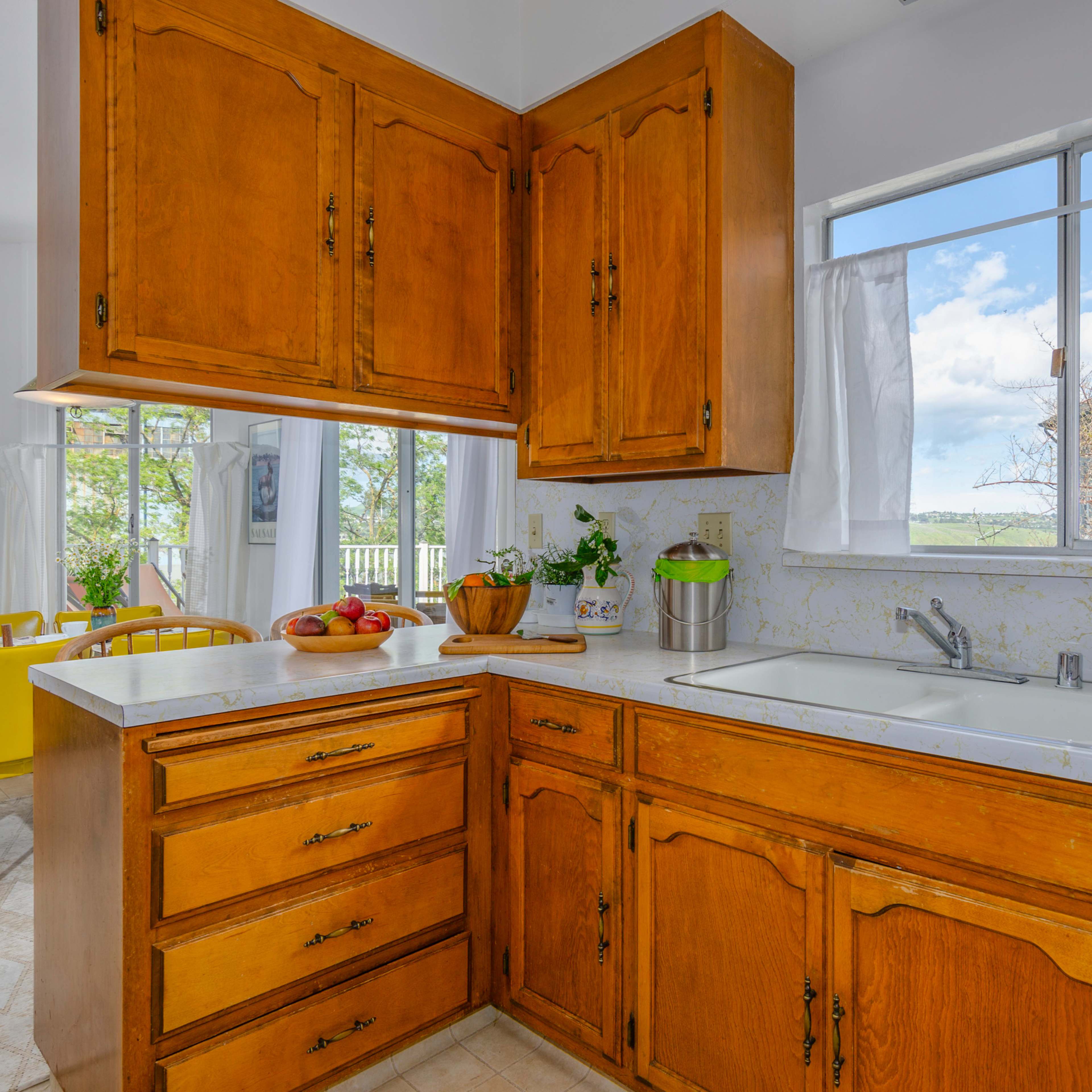 A kitchen with wooden cabinets, a white countertop, and a window overlooking a garden area.