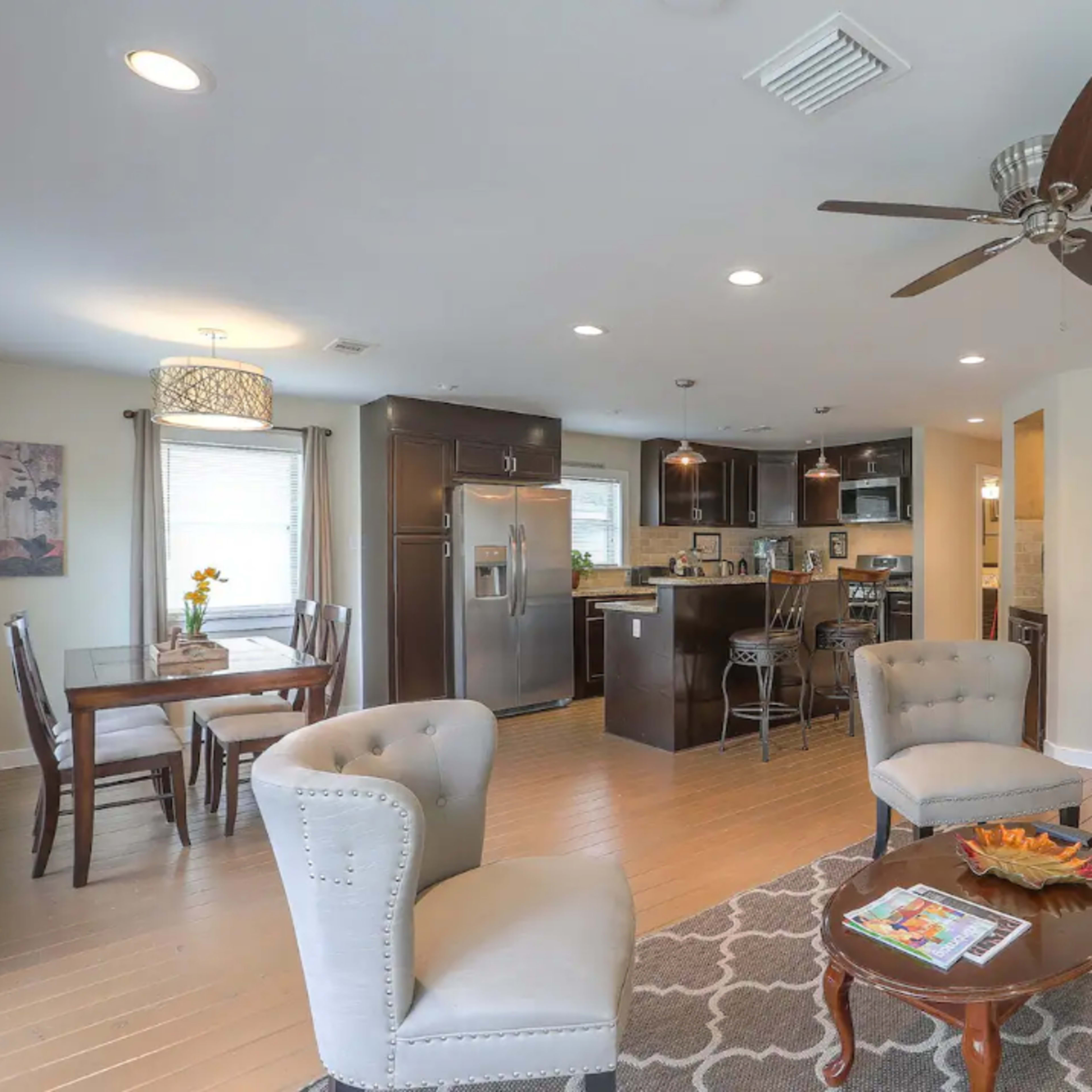 A modern open-concept living space featuring a kitchen, dining area, and seating area with a ceiling fan and a large flat-screen TV.