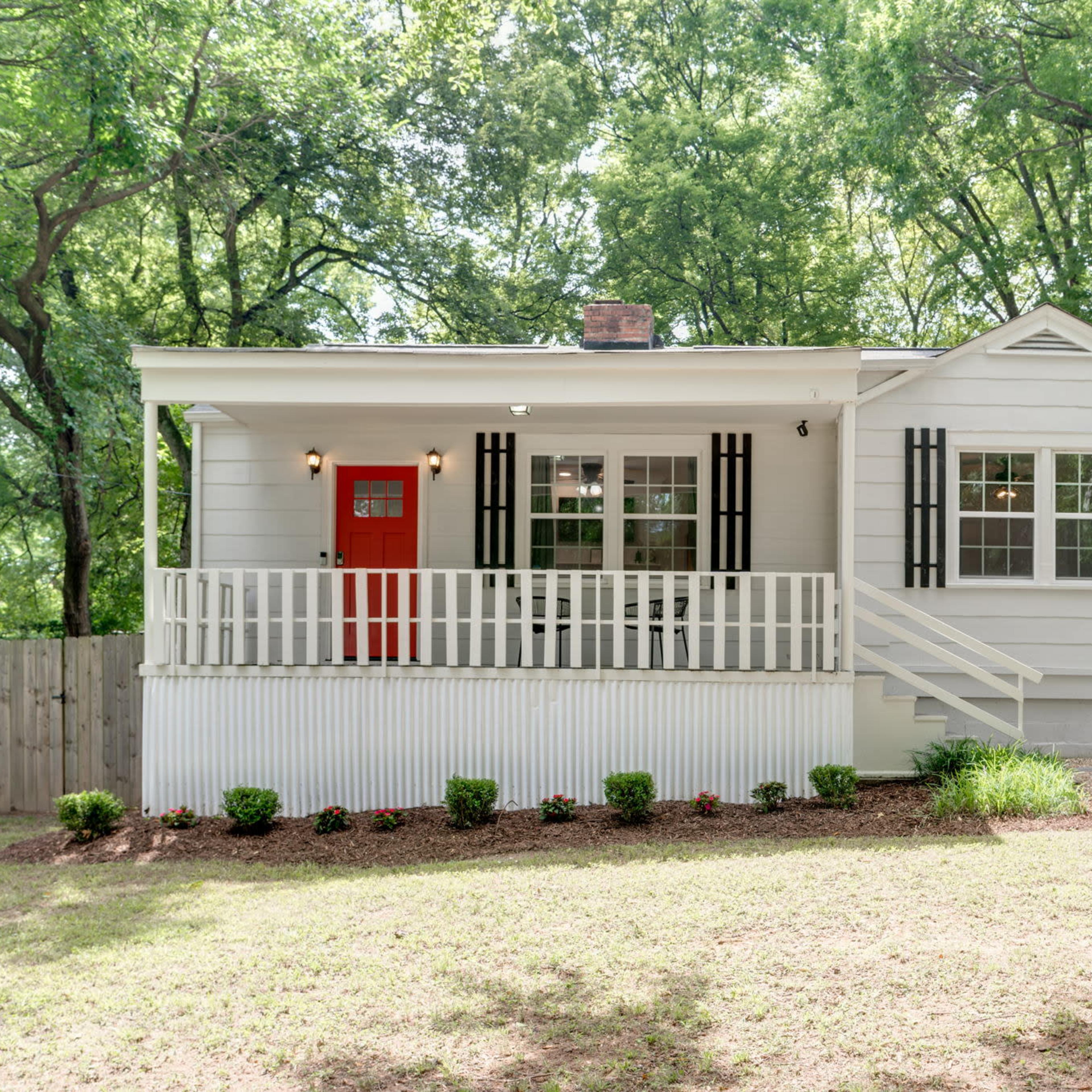 A single-story, white house with a red front door and a porch is set in a grassy area surrounded by trees.